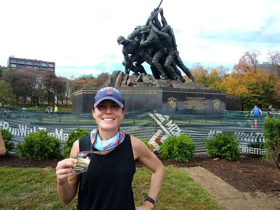 Woman wearing Boston Red Sox hat and black tank top poses with marathon medal in front of a World War 2 memorial