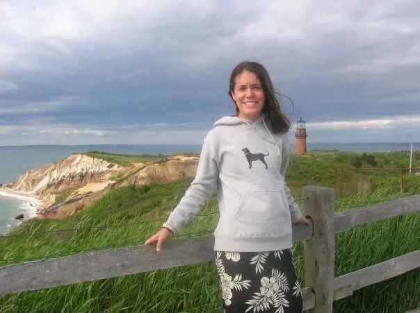 Woman wearing a grey sweatshirt with a black dog on it and a black and white flower print skirt poses in front of a lighthouse and bluffs