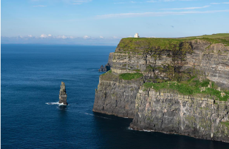 Cliffs overlooking the Atlantic Ocean with a small white lighthouse on top of the cliff and a sea stack jutting out of the water.
