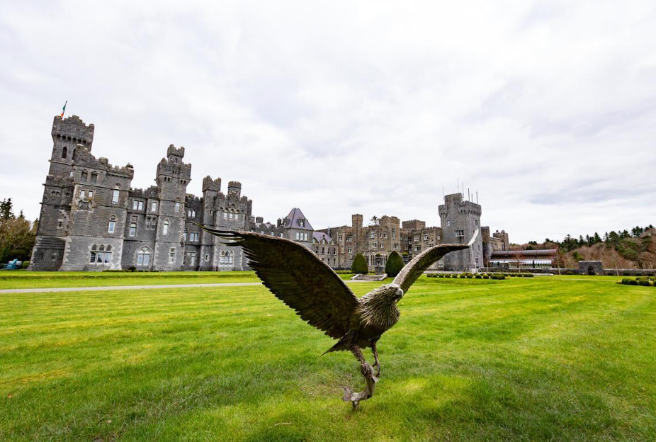 A castle with gray stone walls and towers, surrounded by well-maintained green lawns, with a large statue of a bird in the foreground.
