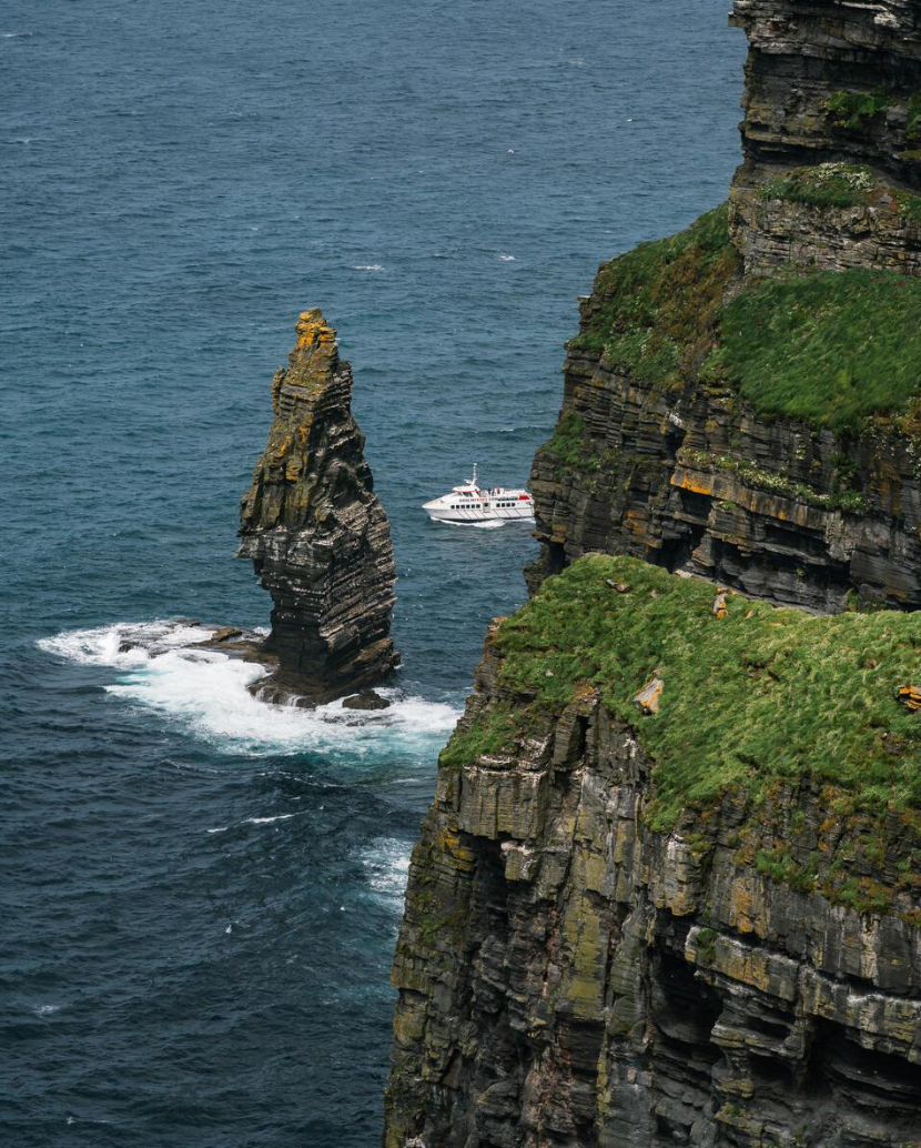 Cliffs with green vegetation overlooking the ocean, with a large rock formation extending into the water and a white boat navigating near the rocks.