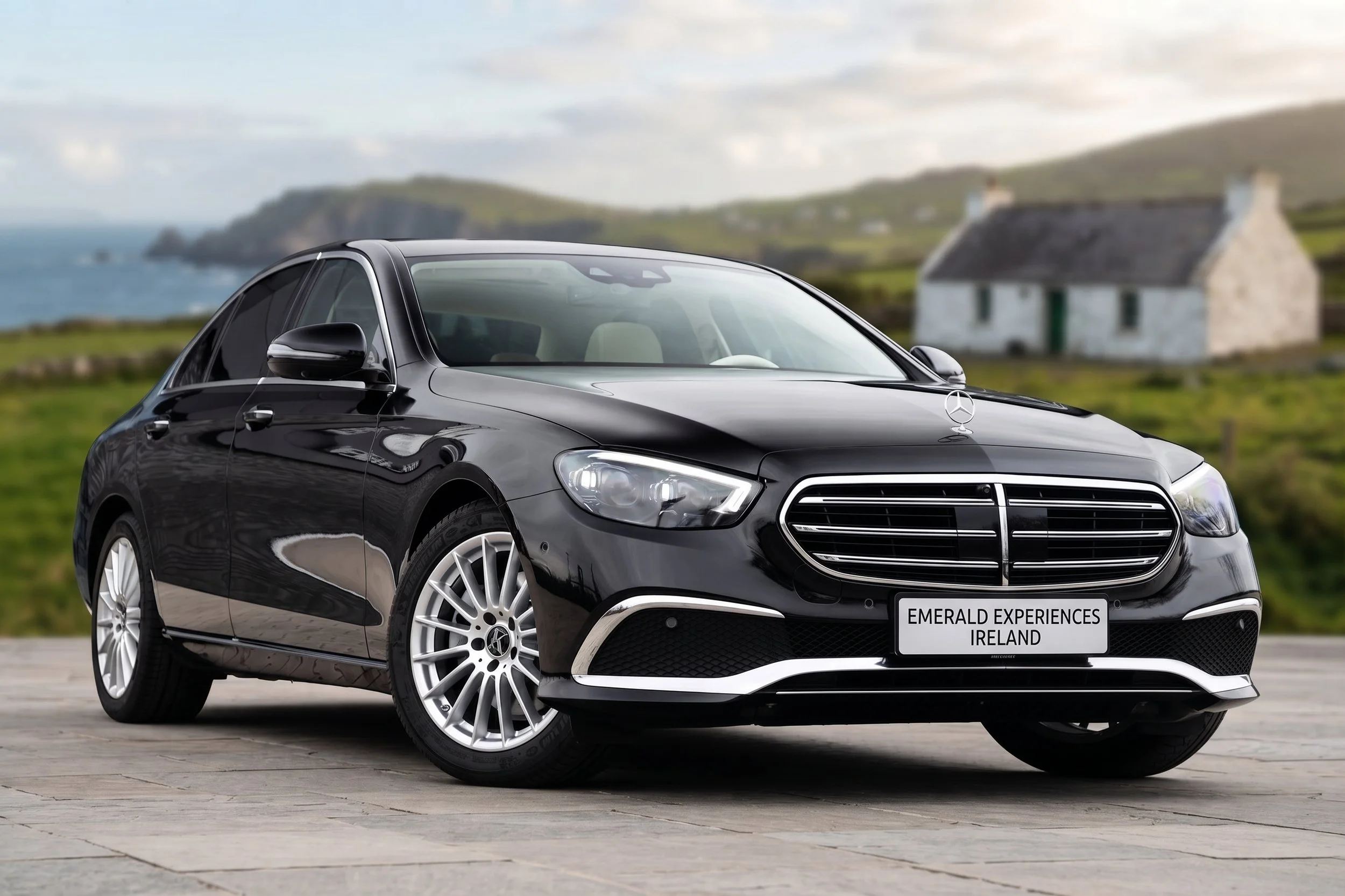 Black Mercedes-Benz car parked outdoors with a rural Irish countryside background, including green hills, a white house, and a cloudy sky.