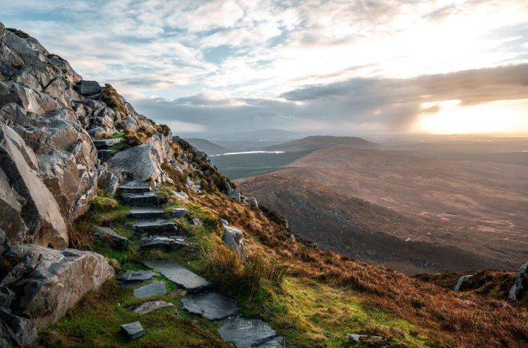 Scenic mountain landscape with stone steps leading up a rocky hillside, overlooking rolling hills, a lake, and a cloudy sky during sunset.