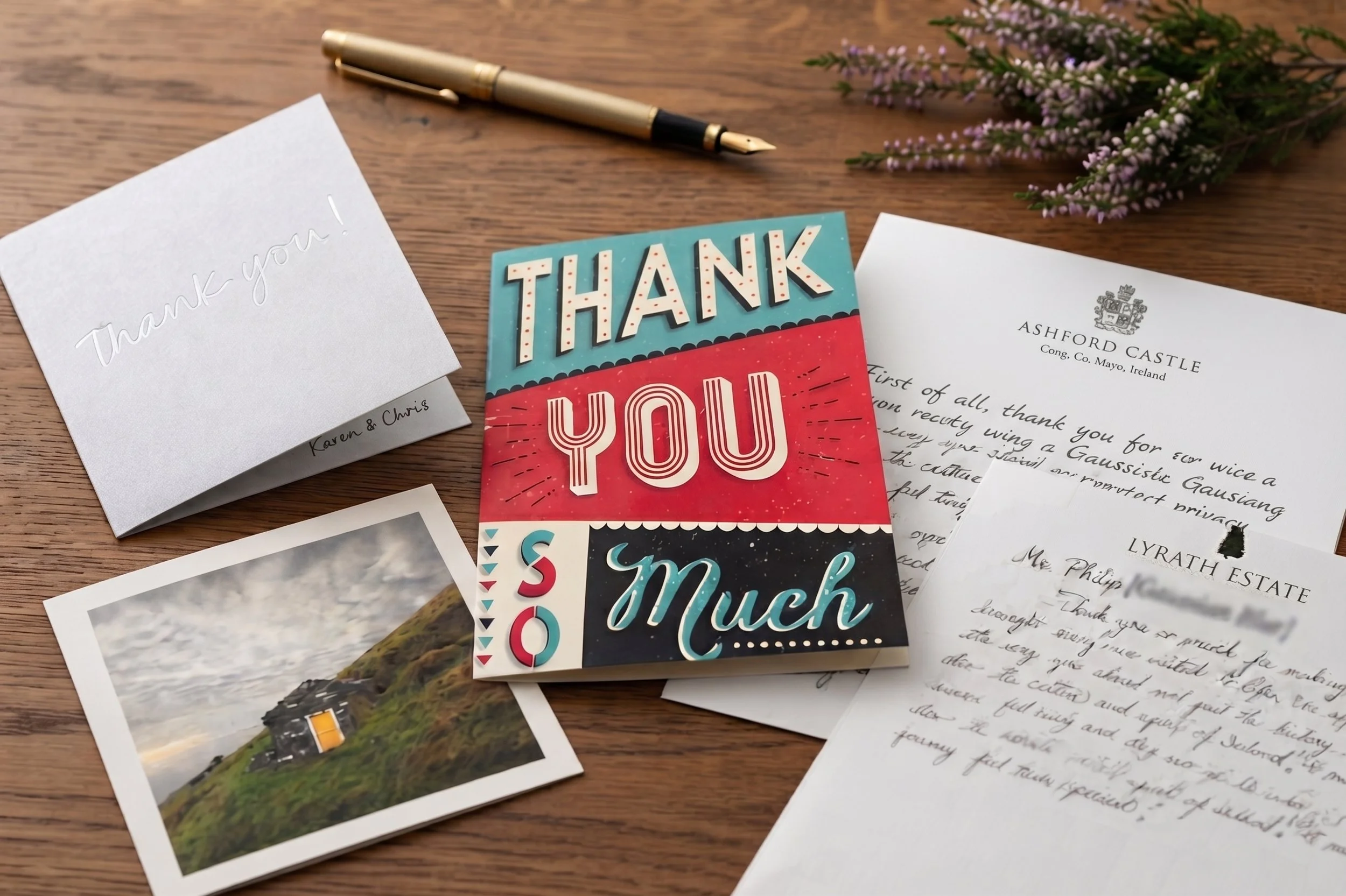 A wooden table with a gold fountain pen, a small bouquet of purple flowers, a white thank you card with cursive writing, colorful thank you greeting cards, and postcards with landscape photos.