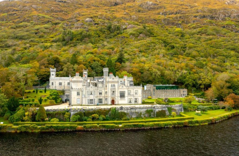 A large, historic castle with multiple towers and turrets, surrounded by lush green gardens, situated on the edge of a lake with a forested hillside in the background.