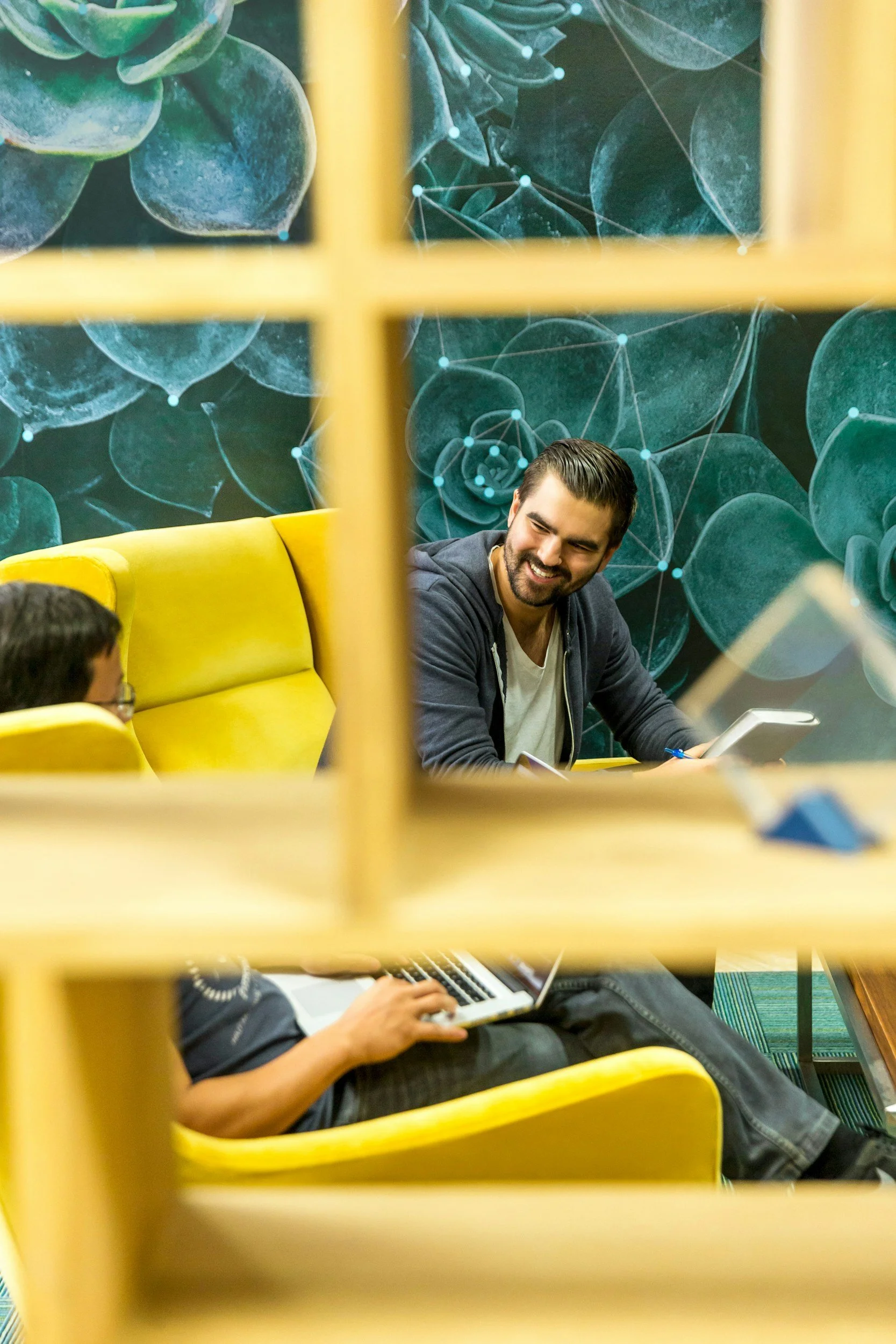 Two men sitting on yellow couches having a conversation, with a decorative dark green floral wall behind them, seen through a yellow grid structure.