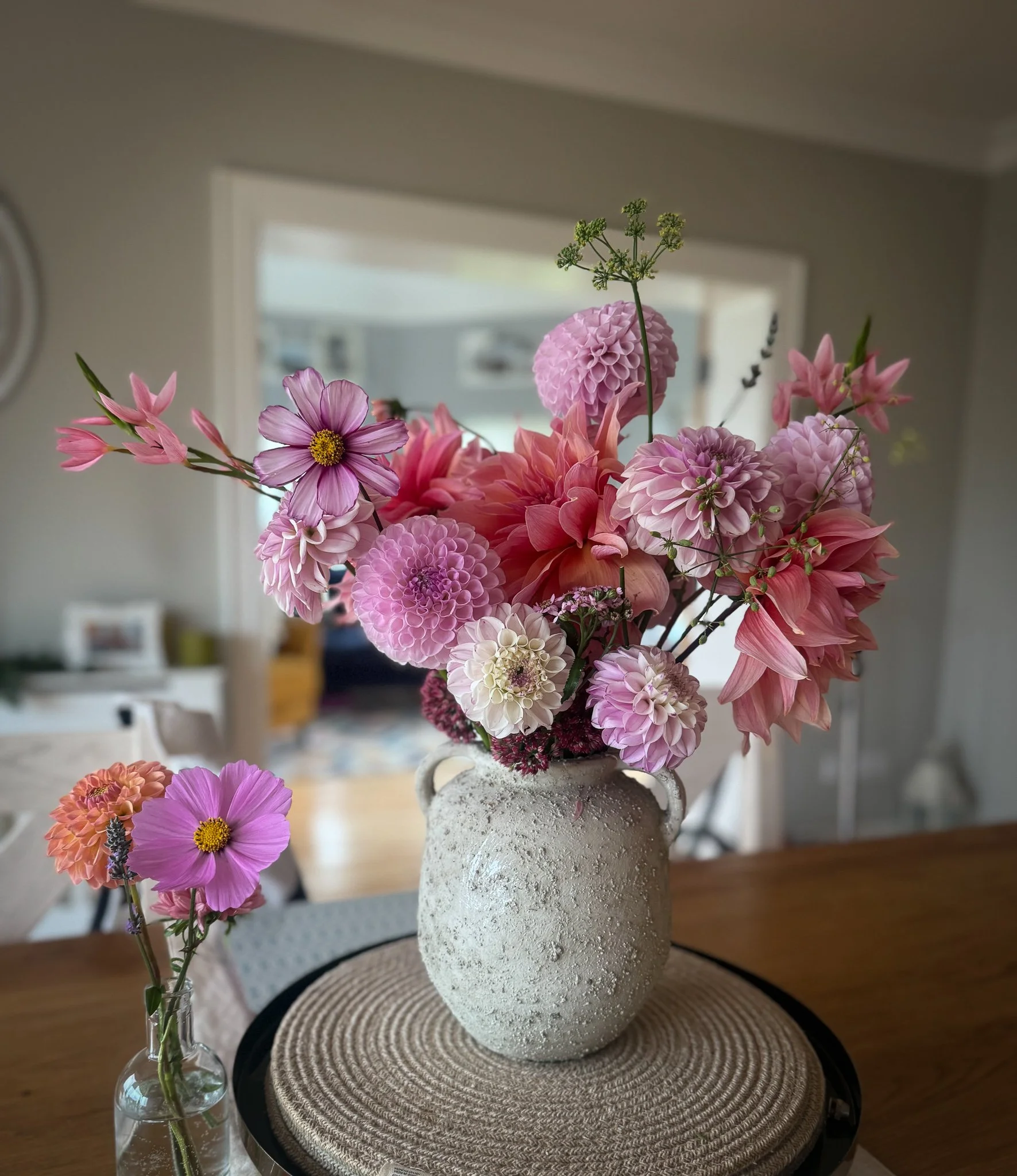 Pink and purple flowers arranged in a textured white ceramic vase on a wooden table, with a smaller flower arrangement in a glass vase nearby.