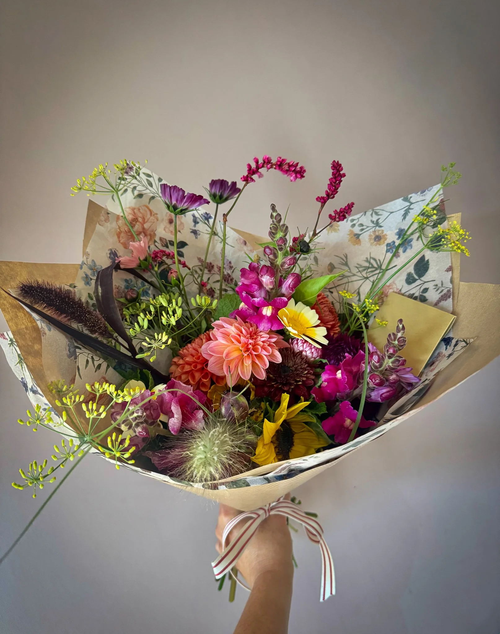 A colorful bouquet of mixed flowers including dahlias, sunflowers, and snapdragons, wrapped in decorative paper and tied with a striped ribbon.