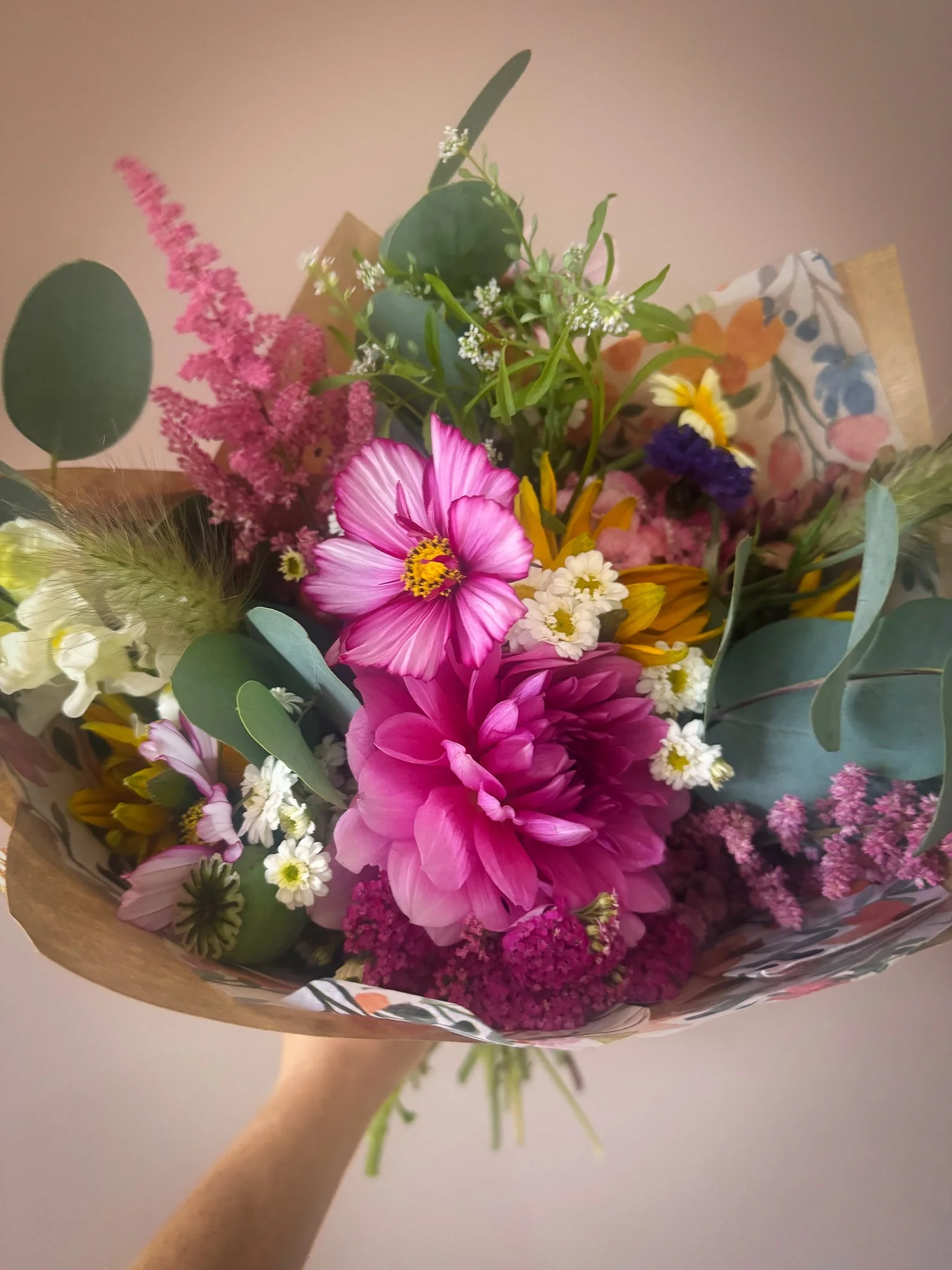 A colorful bouquet of various flowers including pink, purple, yellow, and white blossoms, surrounded by green leaves, wrapped in decorative paper.