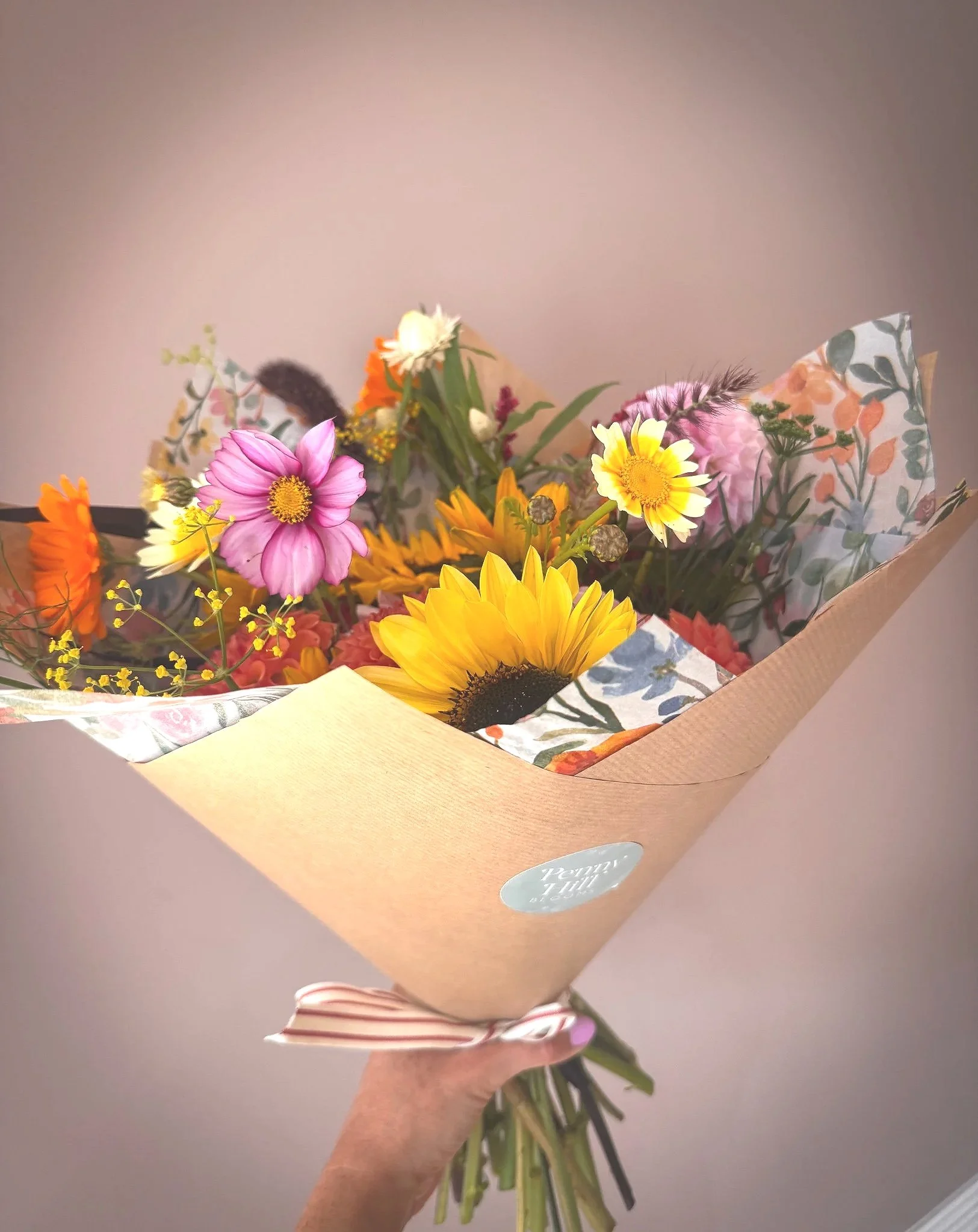 A hand holding a bouquet of colorful flowers wrapped in patterned paper and brown paper, with a small white sticker on the wrapping.