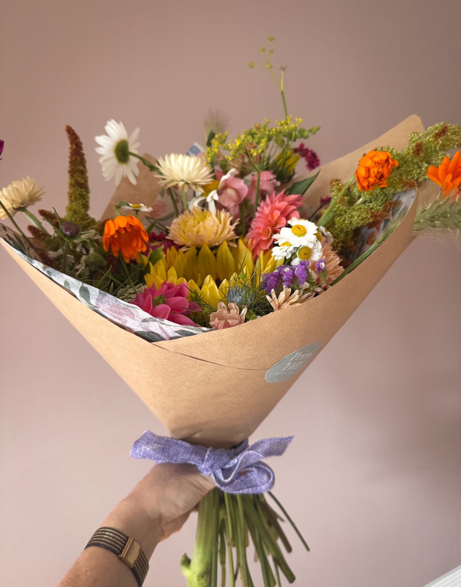 Hand holding a colorful bouquet of mixed flowers wrapped in brown paper with a purple ribbon