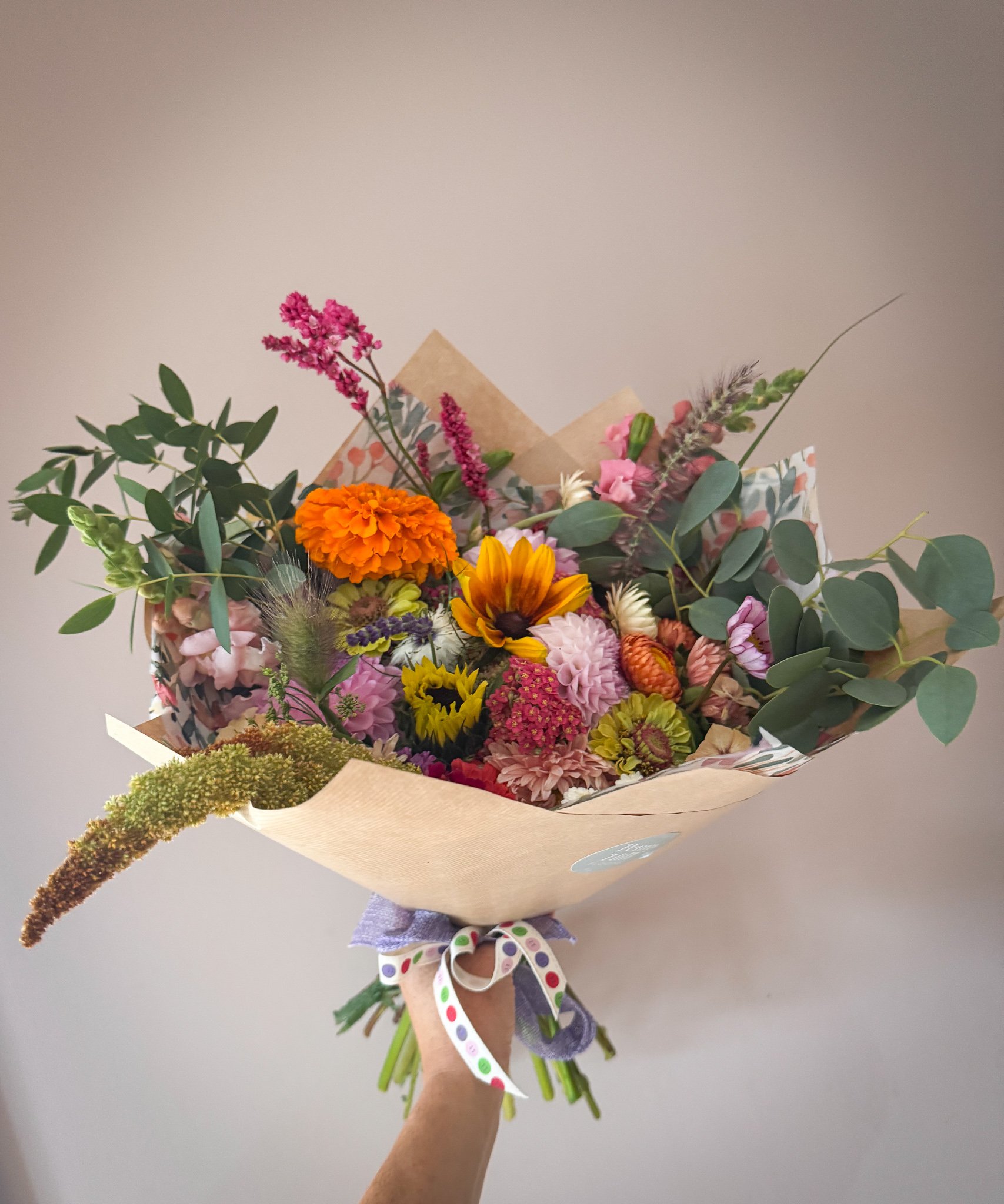 Colorful bouquet of various flowers including marigold, sunflower, pink and purple blooms, green foliage, and wrapped in brown paper with a ribbon.