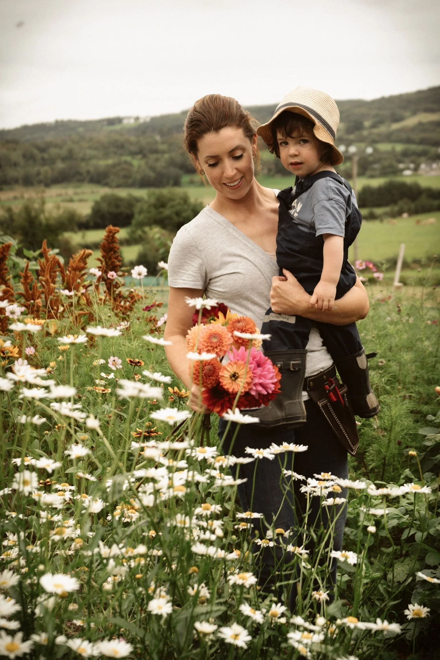A woman holding a young child in a flower garden, with rolling green hills in the background.