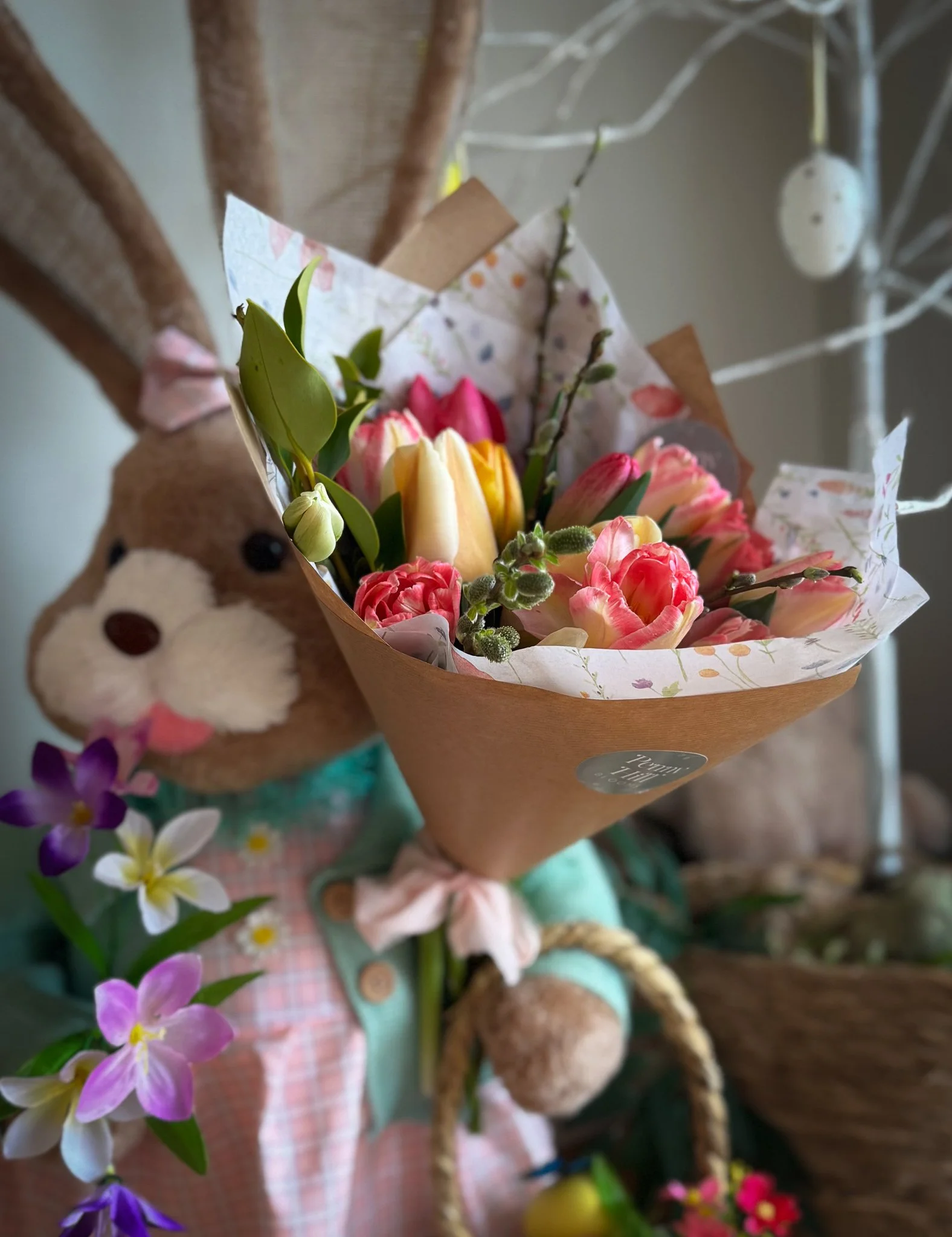 A stuffed animal holding a bouquet of pink, yellow, and white tulips and other flowers, with purple and white flowers nearby. The background features a white decorative branch and hanging white ornaments.