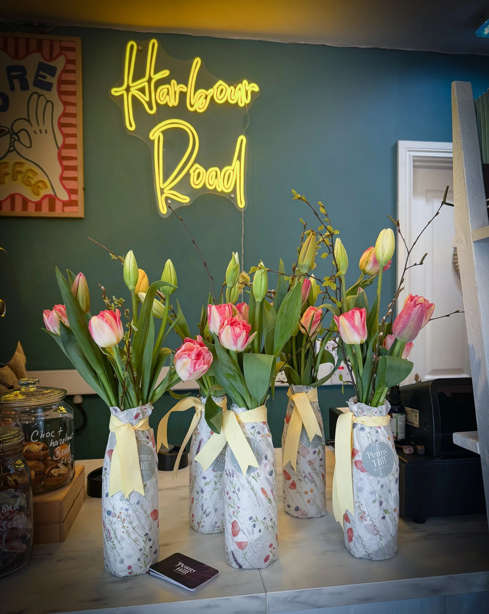 Five vases with pink and white tulips wrapped in decorative paper with yellow ribbons on a marble countertop, with a neon sign reading "Harlow Road" in yellow above.