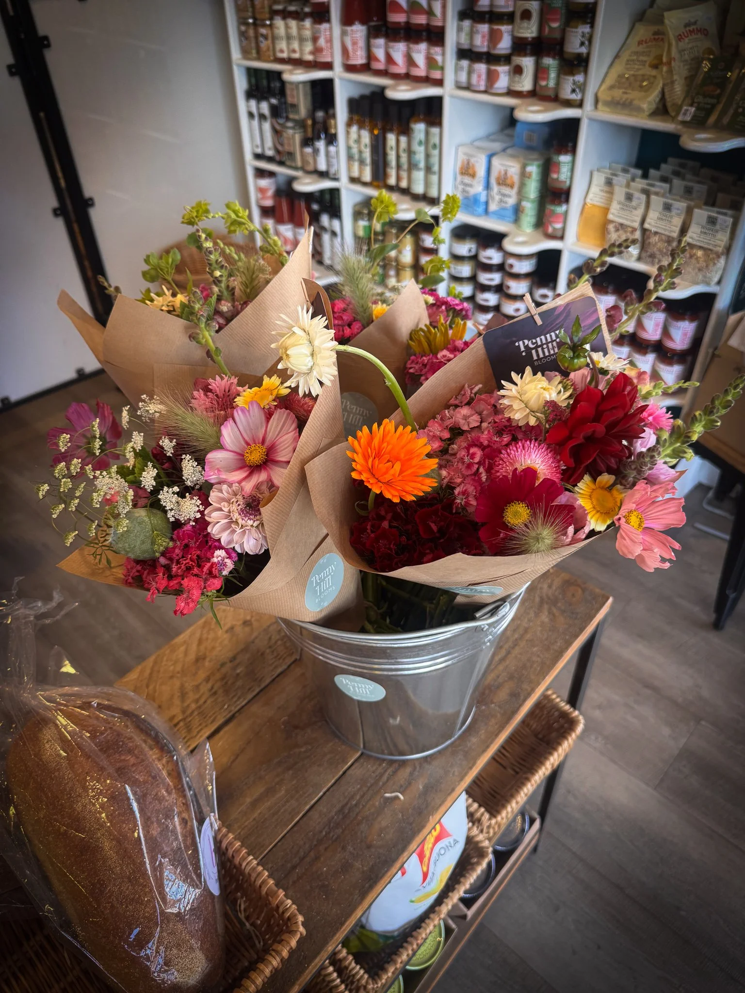 Colorful flower bouquets wrapped in brown paper in a metal bucket on a wooden table inside a store.