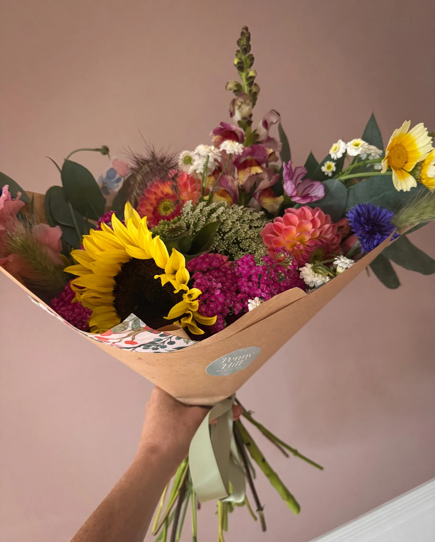 A person holding a colorful bouquet of fresh flowers wrapped in brown paper, featuring sunflowers, daisies, pink and purple flowers, and green foliage.