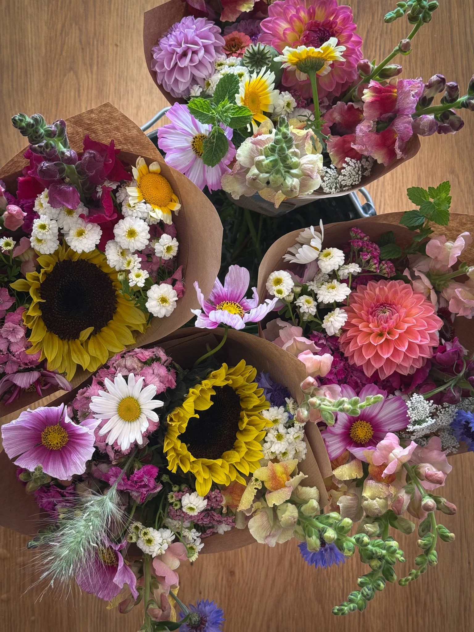 Multiple bouquets of colorful flowers on a wooden surface, including sunflowers, daisies, dahlias, snapdragons, and other mixed wildflowers.