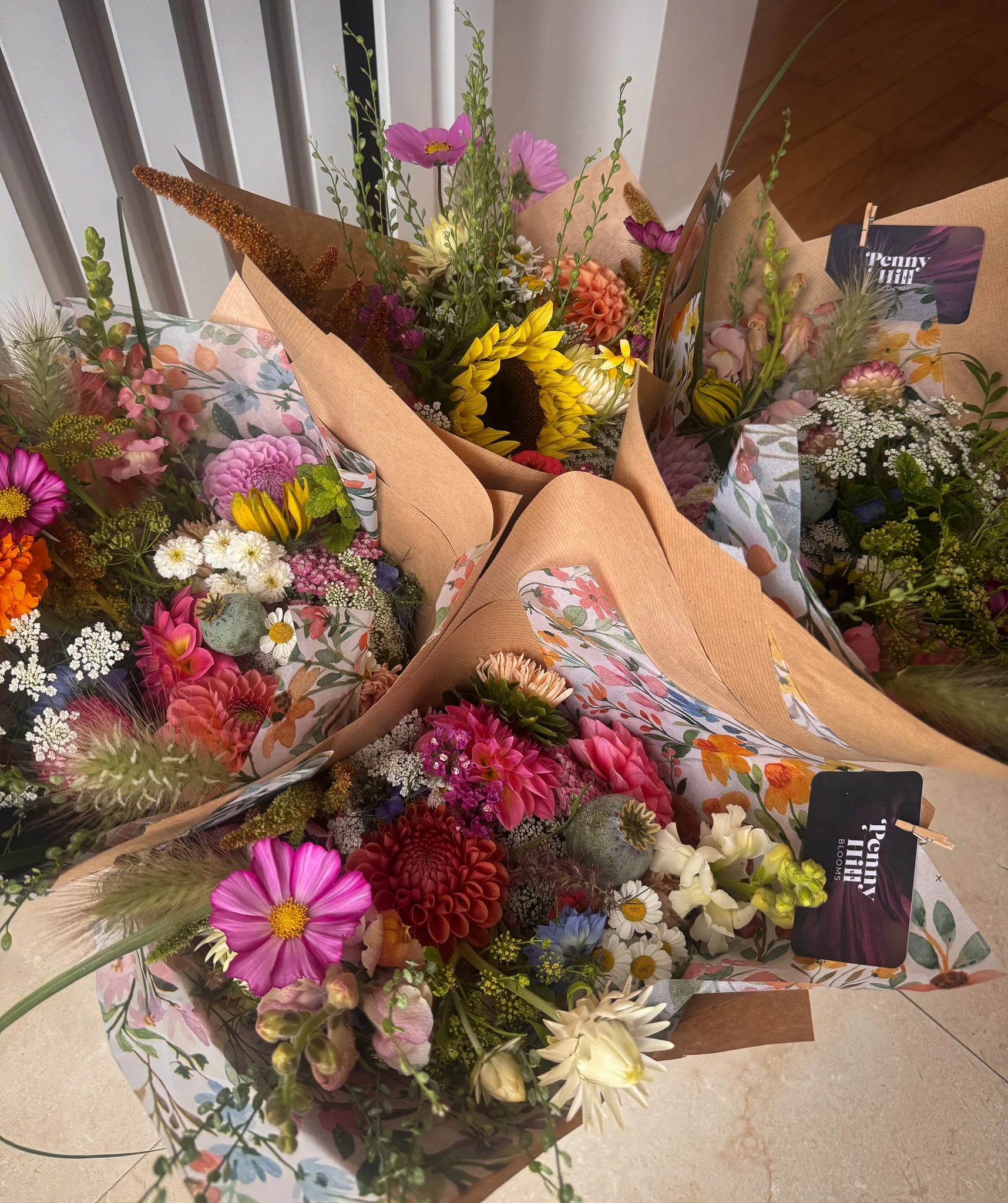 Multiple colorful bouquets of flowers wrapped in brown paper and decorative floral sheets, with a variety of flowers including sunflowers, dahlias, daisies, and others