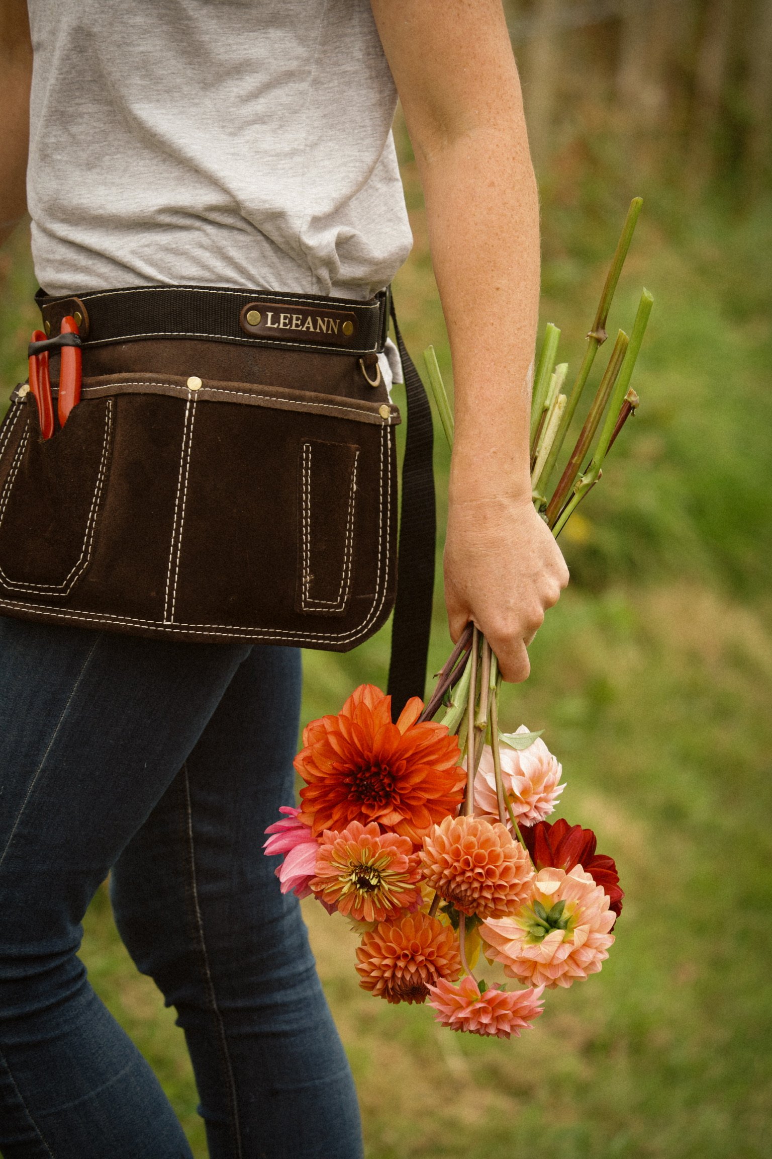 Person holding a bunch of orange and pink flowers, wearing dark jeans and a grey shirt, with a black tool belt.