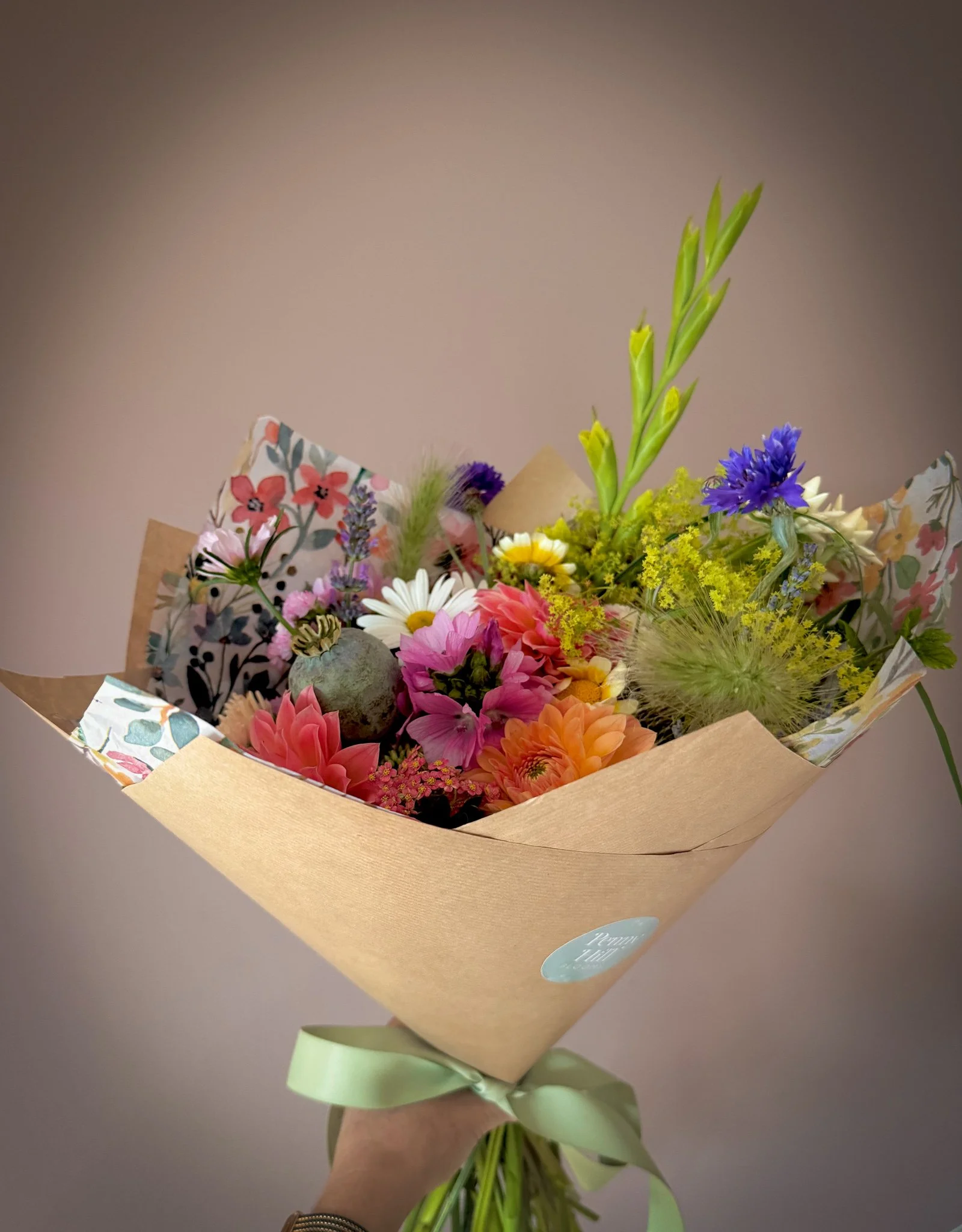 Hand holding a colorful bouquet of flowers wrapped in brown paper and decorative floral paper, tied with a light green ribbon.