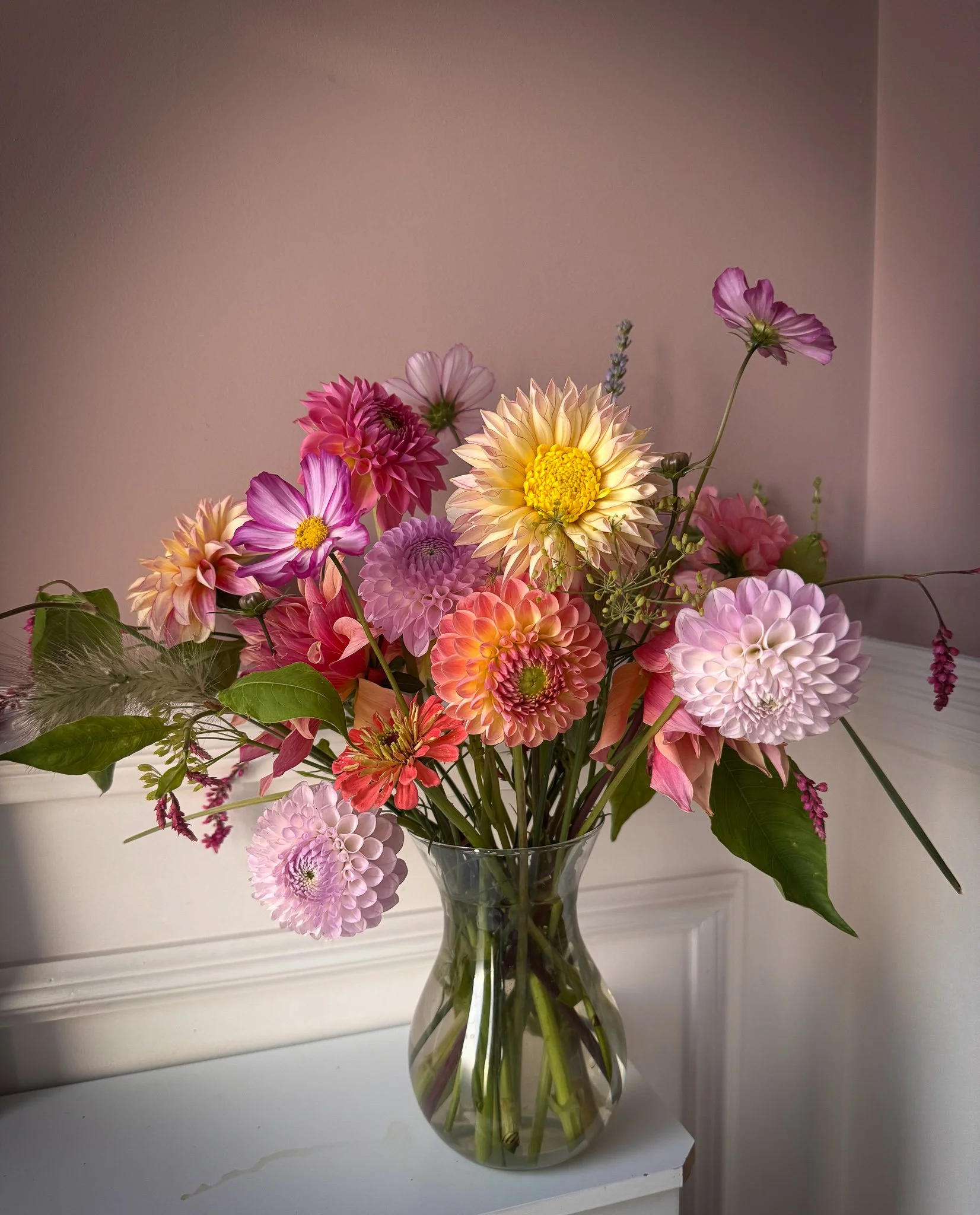 A colorful bouquet of mixed dahlias and other flowers in a clear glass vase on a white surface against a light pink wall.
