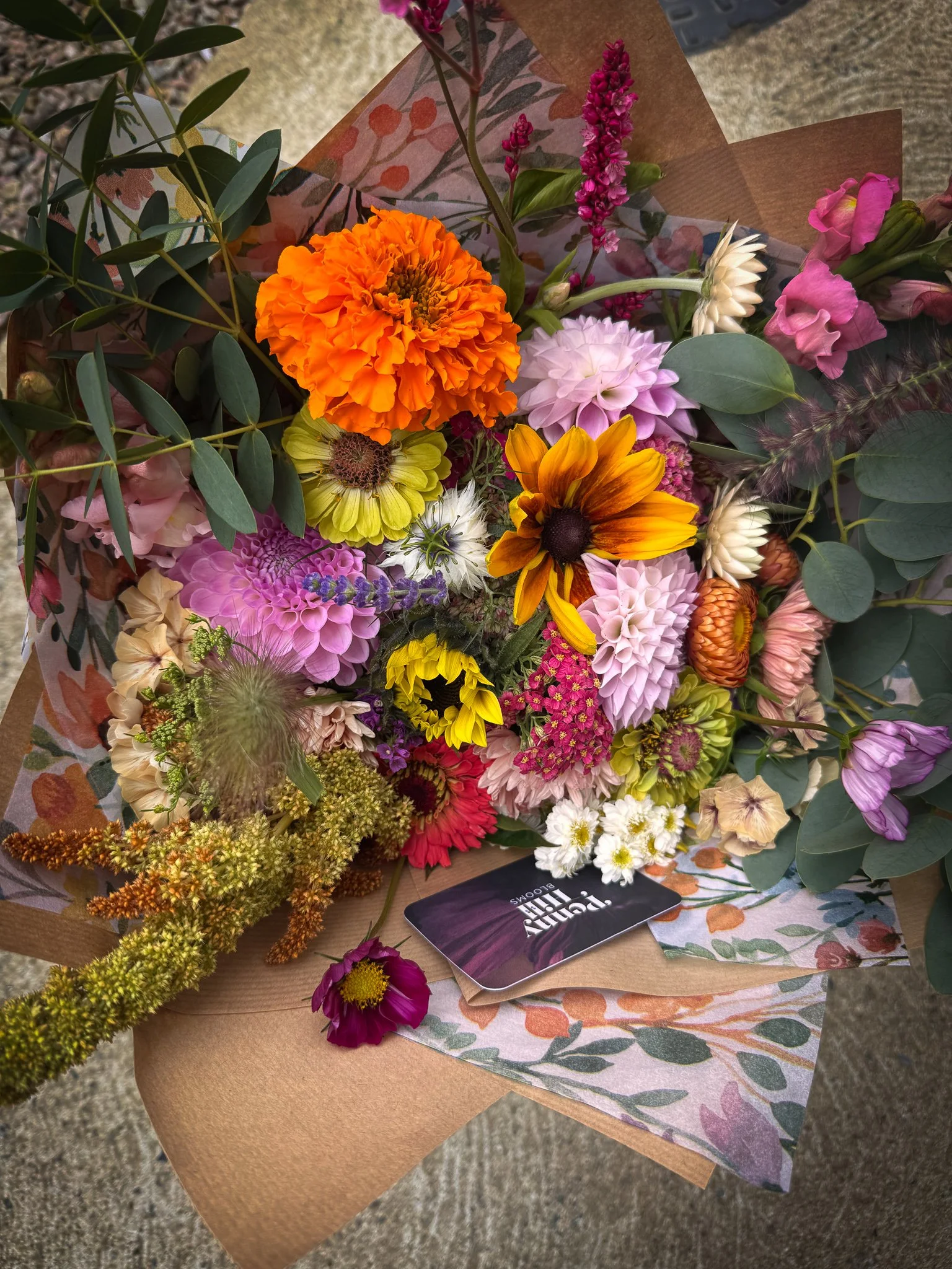 A colorful flower bouquet with marigolds, daisies, dahlias, and various greenery, wrapped in patterned paper.