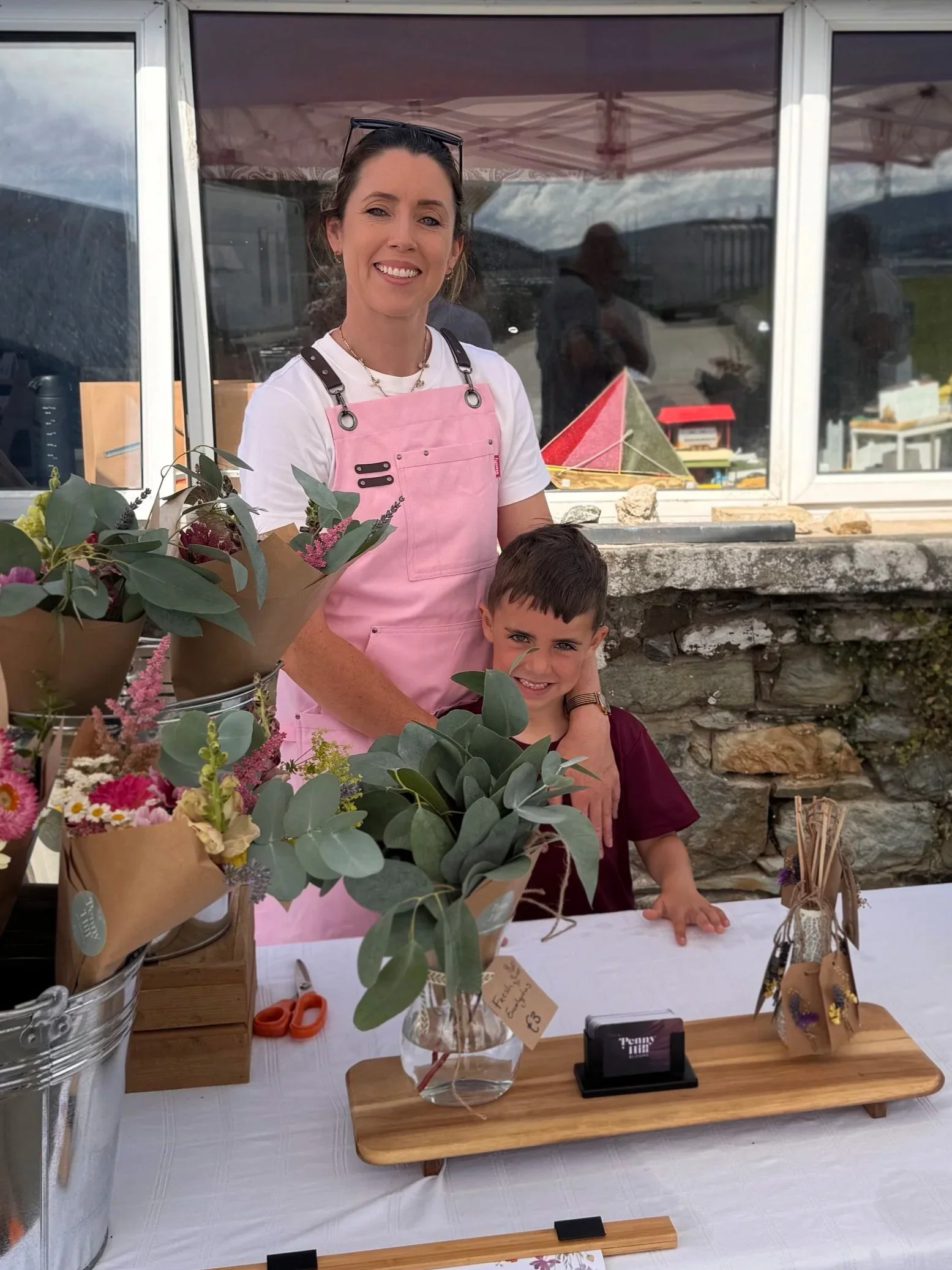A woman in a pink apron and a young boy standing behind a table with bouquets of flowers, a vase, and craft supplies outside a building with large windows.