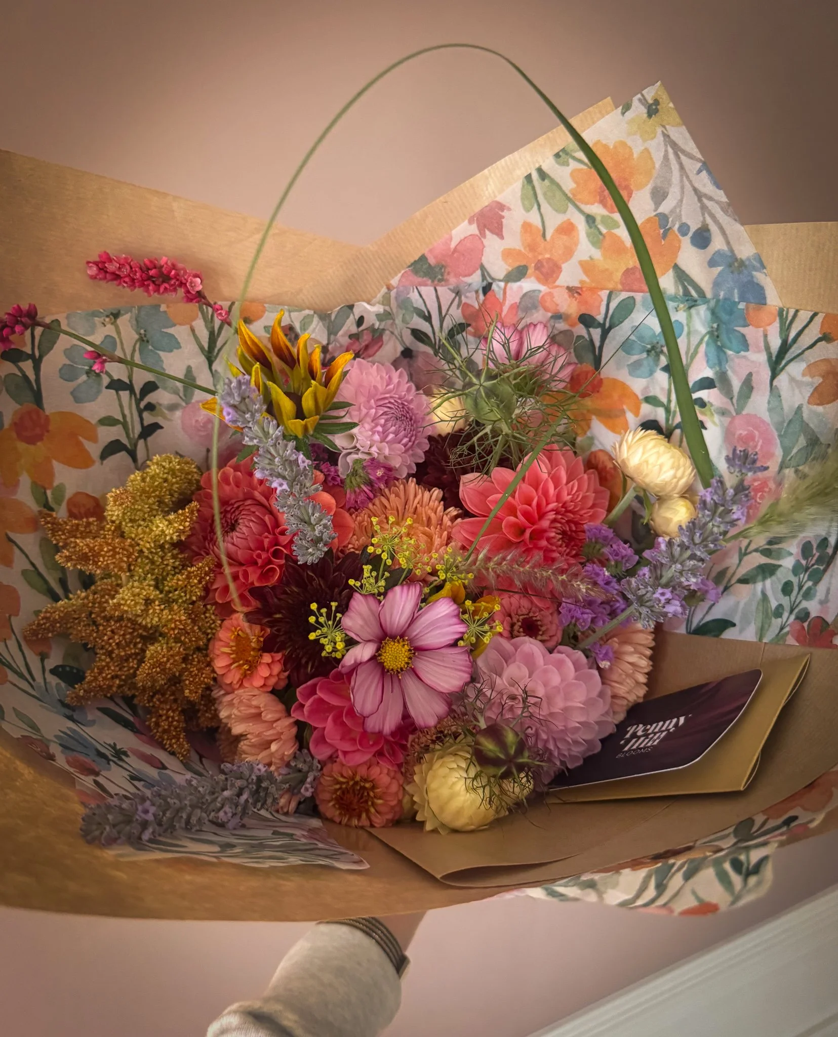 Colorful bouquet of flowers wrapped in floral paper, including pink dahlias, purple lavender, yellow accents, and various greenery.