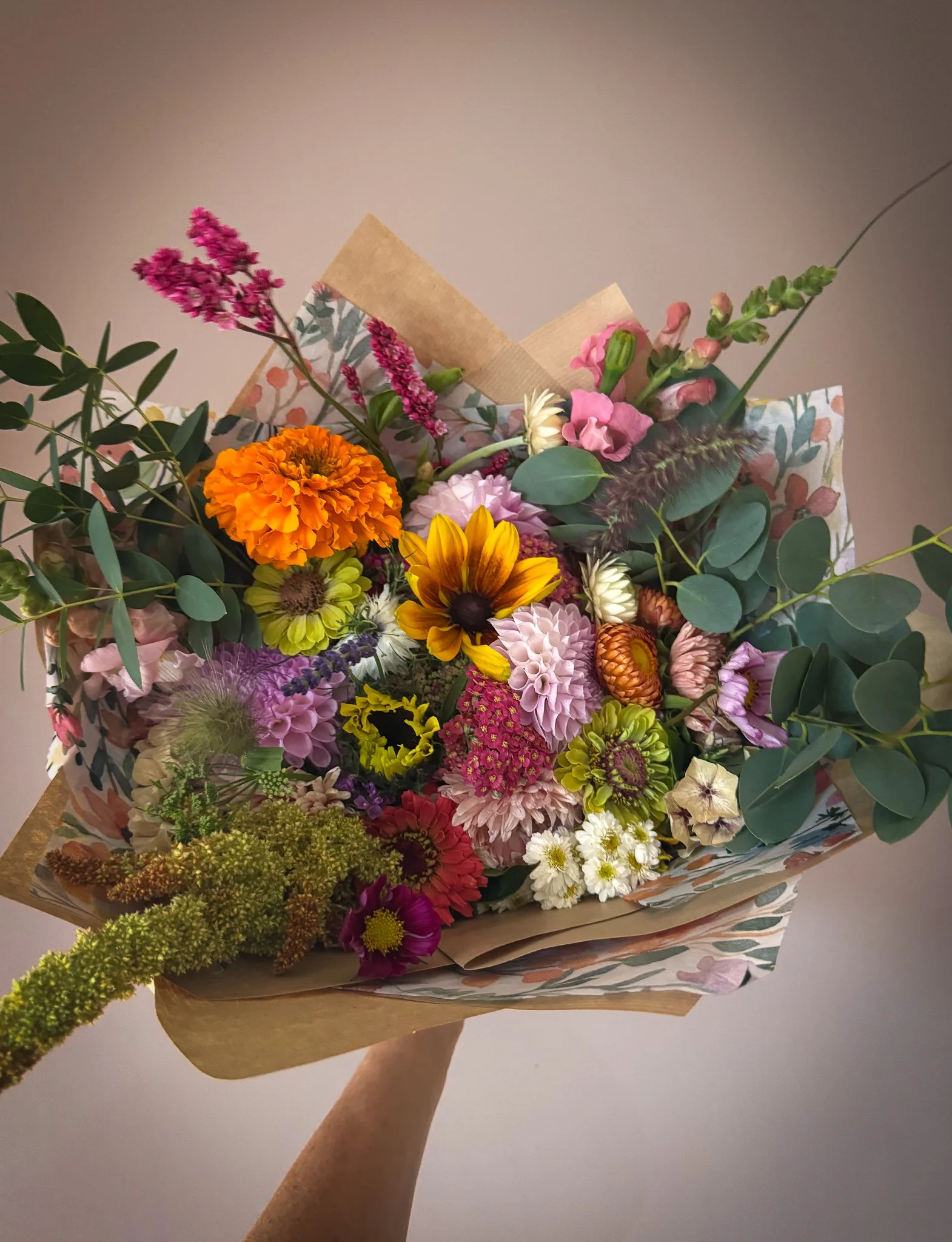 Colorful bouquet of assorted flowers including marigolds, sunflowers, dahlias, lisianthus, and greenery, wrapped in decorative paper.