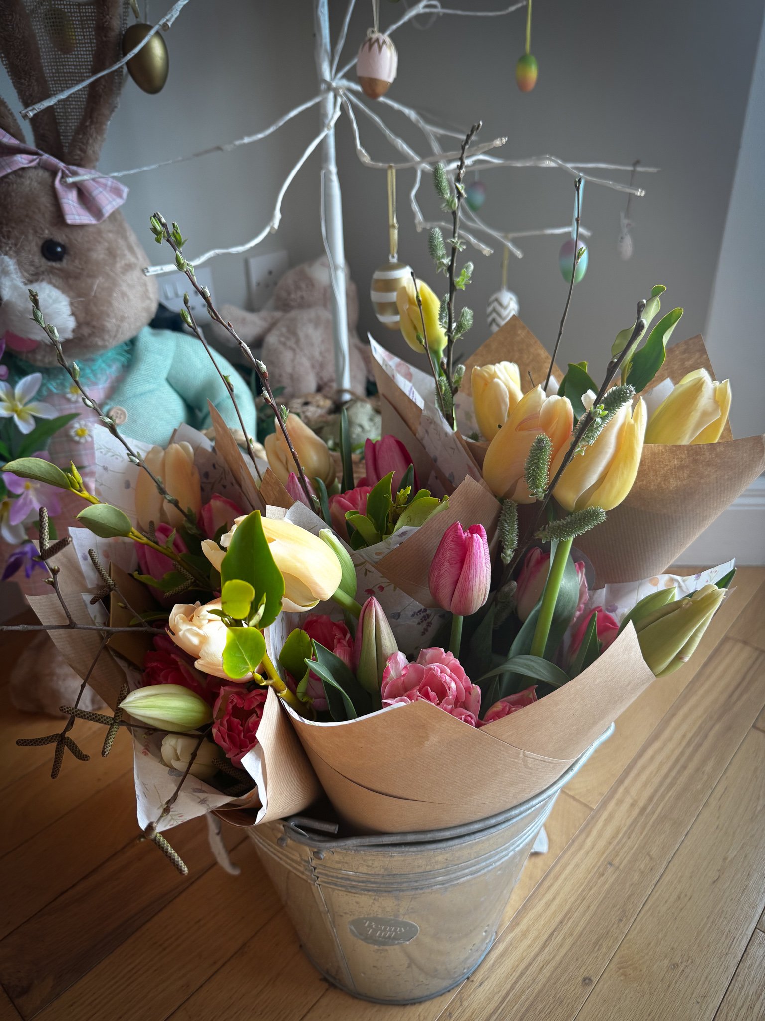 A bouquet of pink and yellow tulips and other flowers in a metal bucket, with wrapped paper, placed on wooden floor. In the background, a plush rabbit toy with a pink bow and a stuffed bear are visible, along with a decorative white tree with hanging