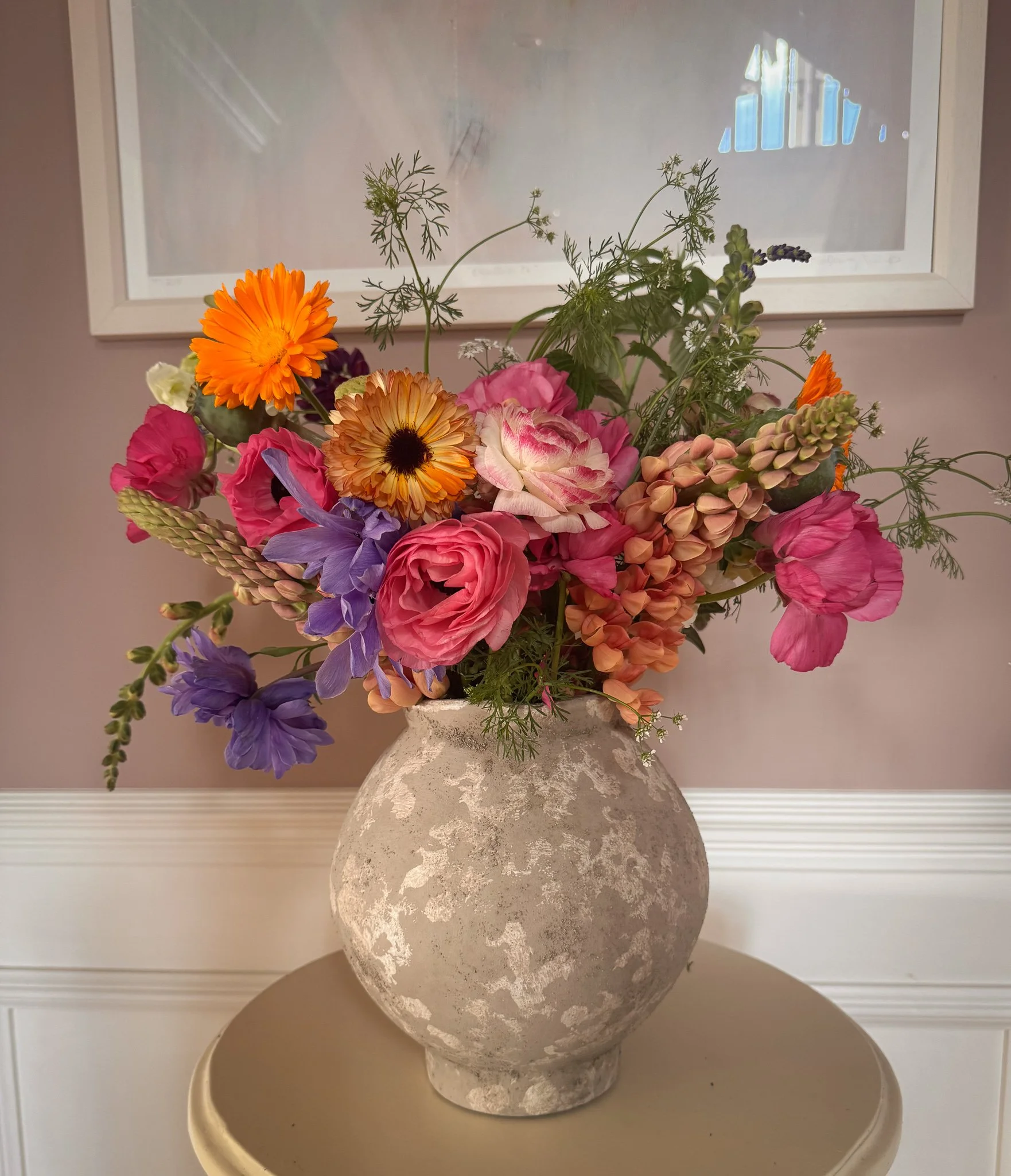 A bouquet of mixed pink, purple, orange, and white flowers in a textured, off-white ceramic vase on a small round table in front of a wall with a framed picture.