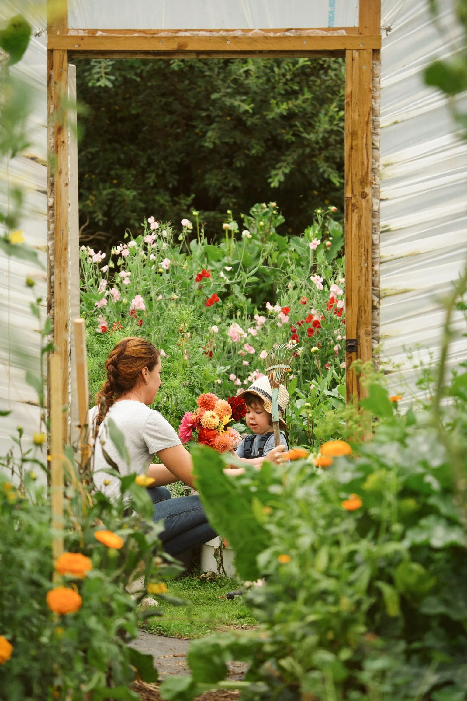 A woman and a young boy, possibly mother and son, sitting in a lush vegetable garden filled with colorful flowers and plants, engaged in a gardening activity.