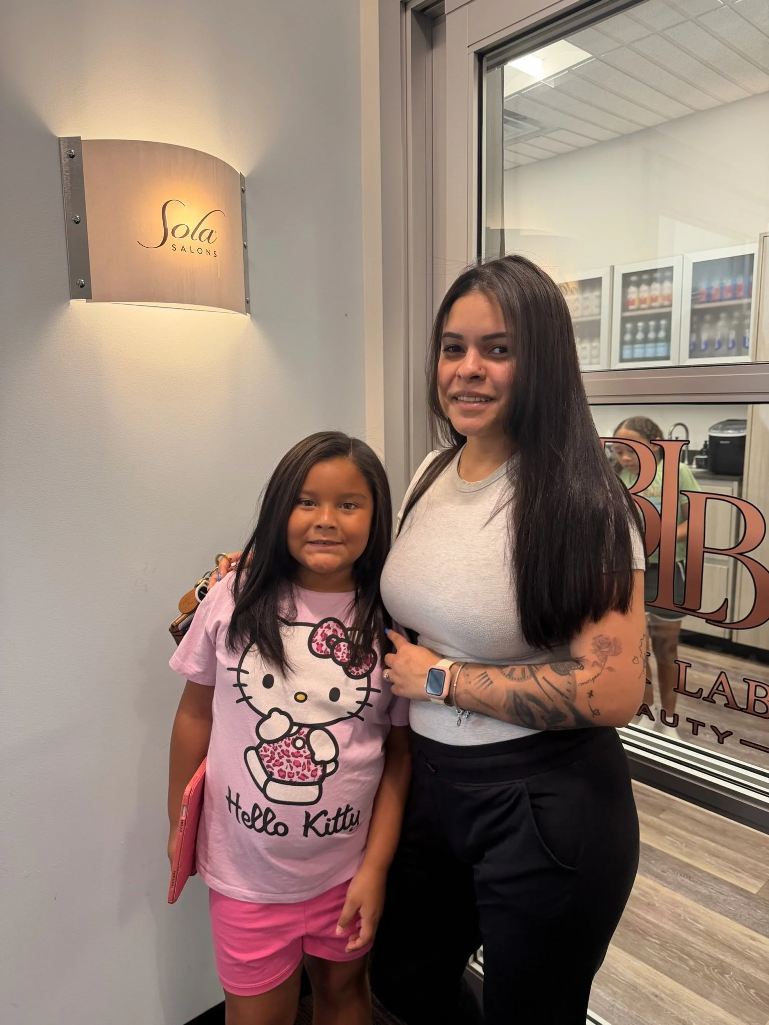 Two women standing together inside a beauty salon, smiling at the camera. One is a young girl wearing a pink Hello Kitty shirt and pink shorts, and the other is an adult woman with long dark hair, tattoos on her arms, and wearing a light-colored t-sh