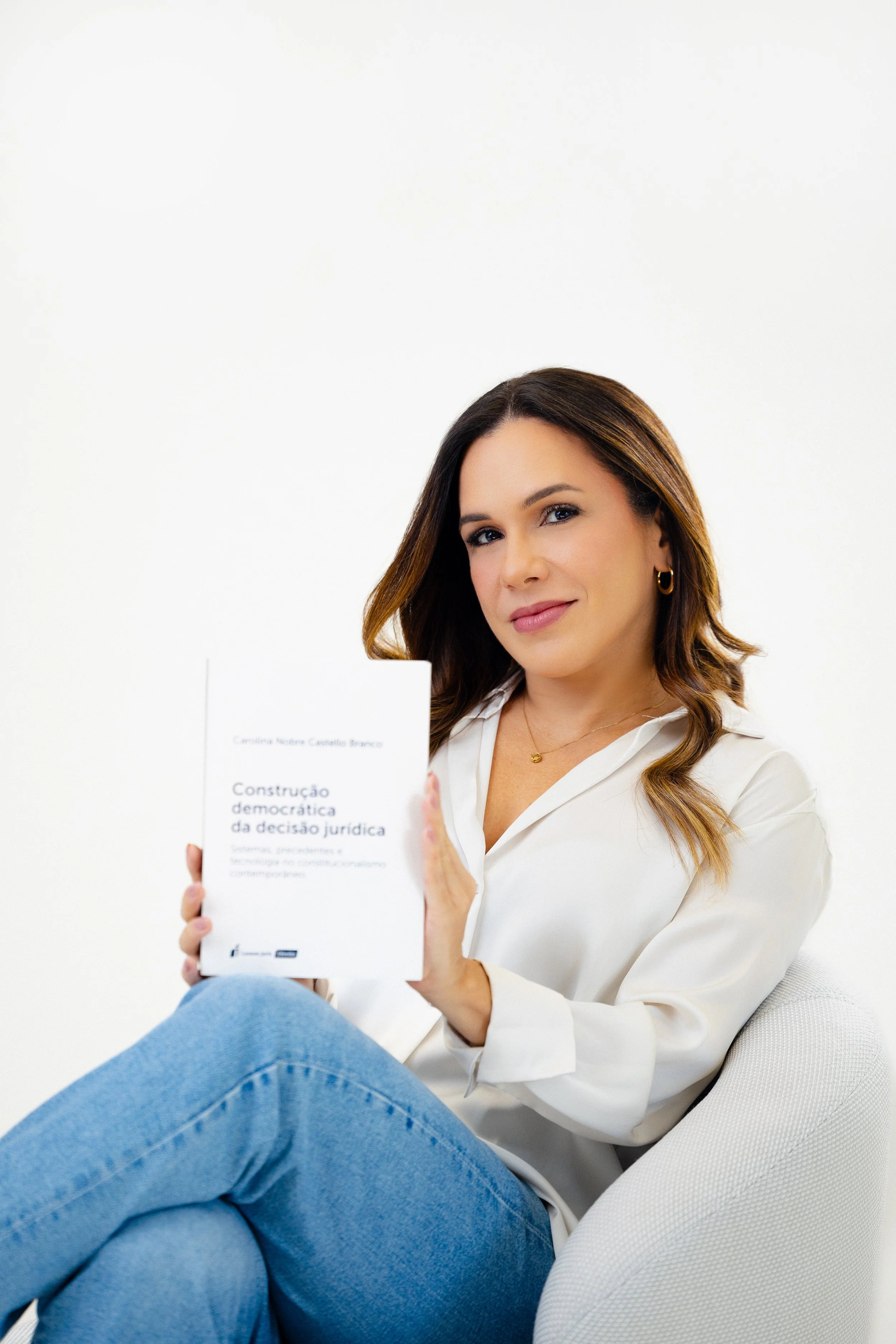 Mulher de cabelo castanho com mechas loiras, vestindo blusa branca e jeans azuis, sentada em uma cadeira branca, segurando um livro com capa branca