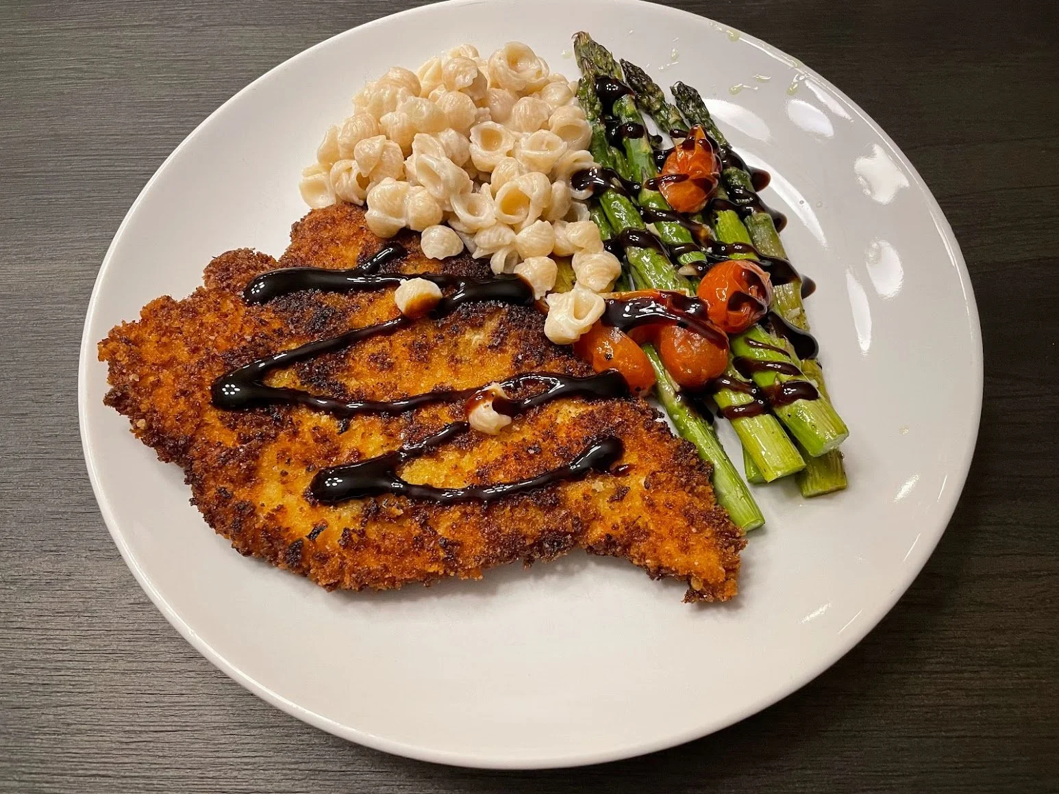 Plate of breaded and fried fish fillet with a drizzle of dark sauce, served with macaroni and cheese, sautéed green asparagus with cherry tomatoes, and garnished with a balsamic glaze.