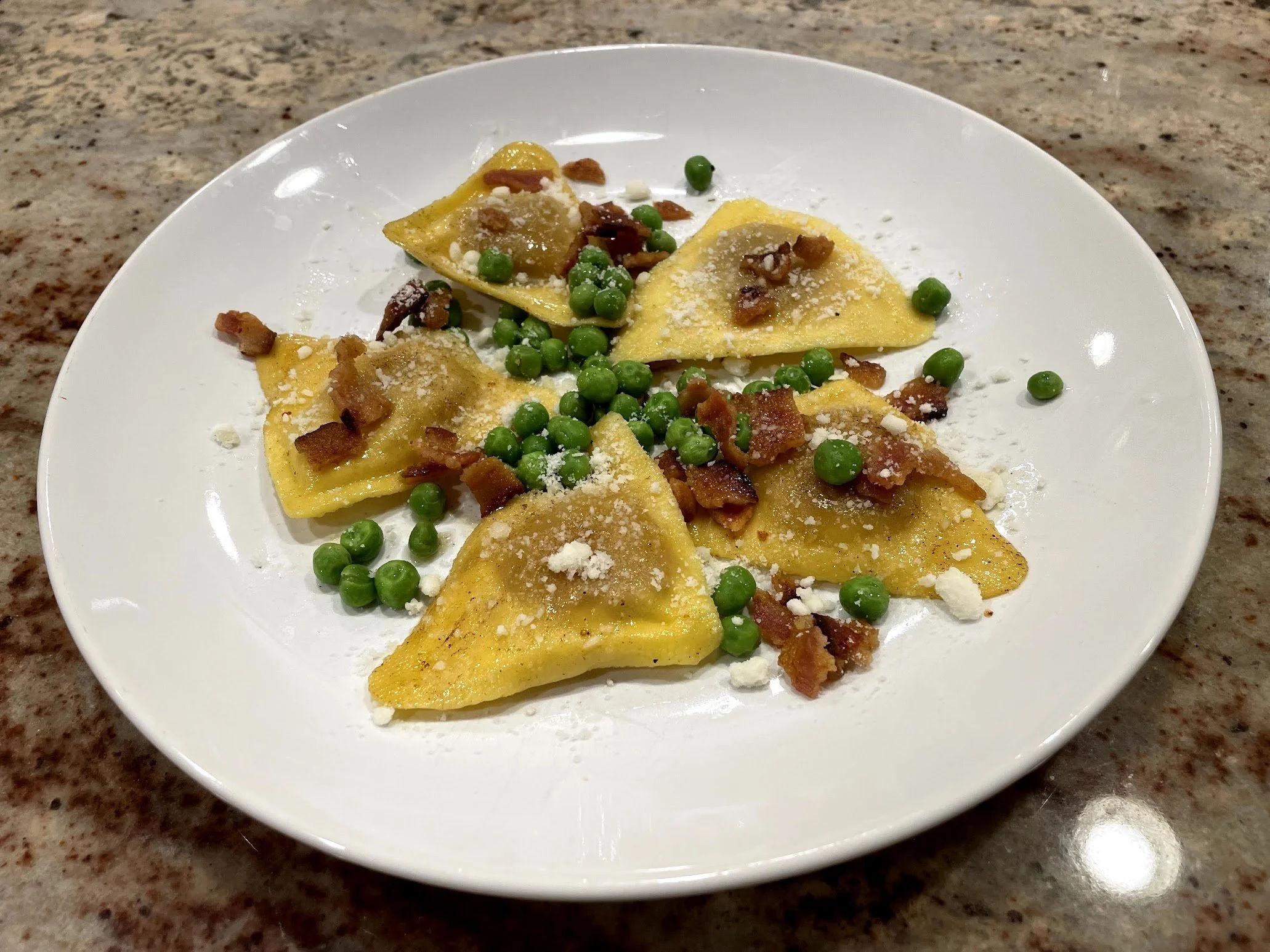 Plate of cooked tortellini pasta with green peas, bacon bits, and grated cheese, on a marble countertop.