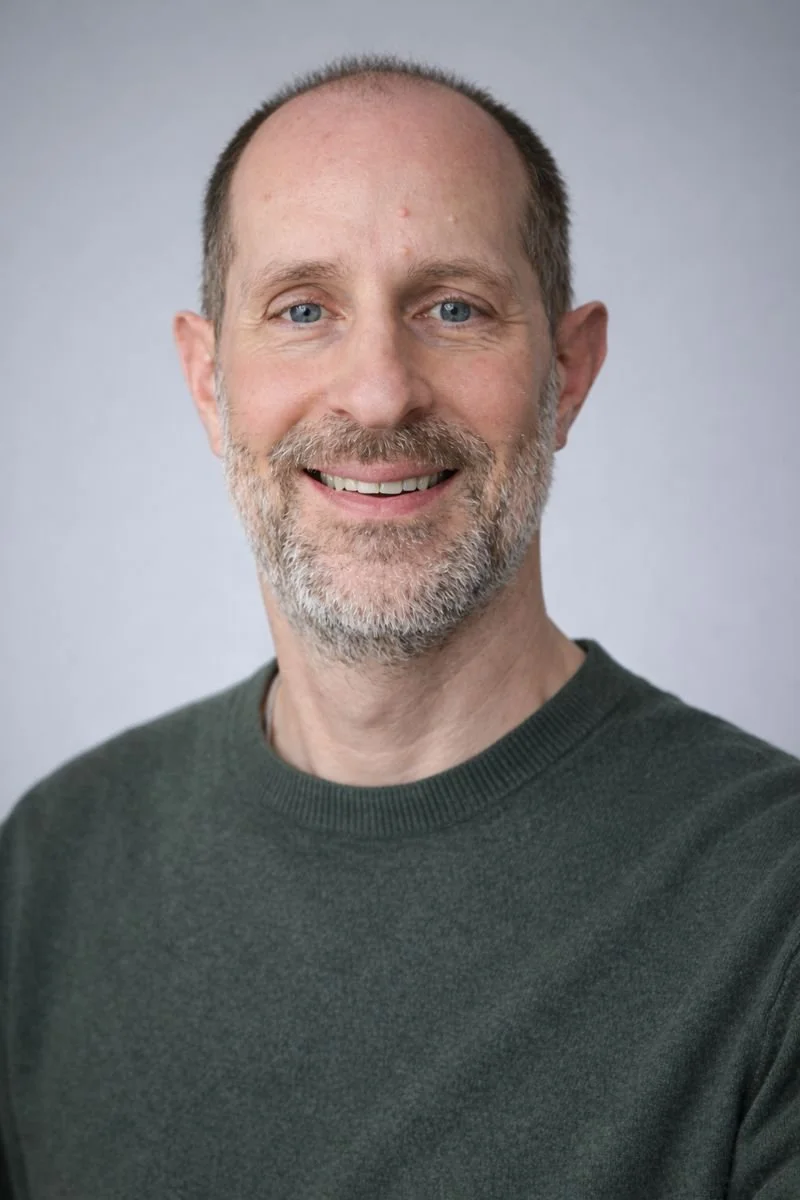 A middle-aged man with a beard, smiling, wearing a dark green sweater, with a plain gray background.