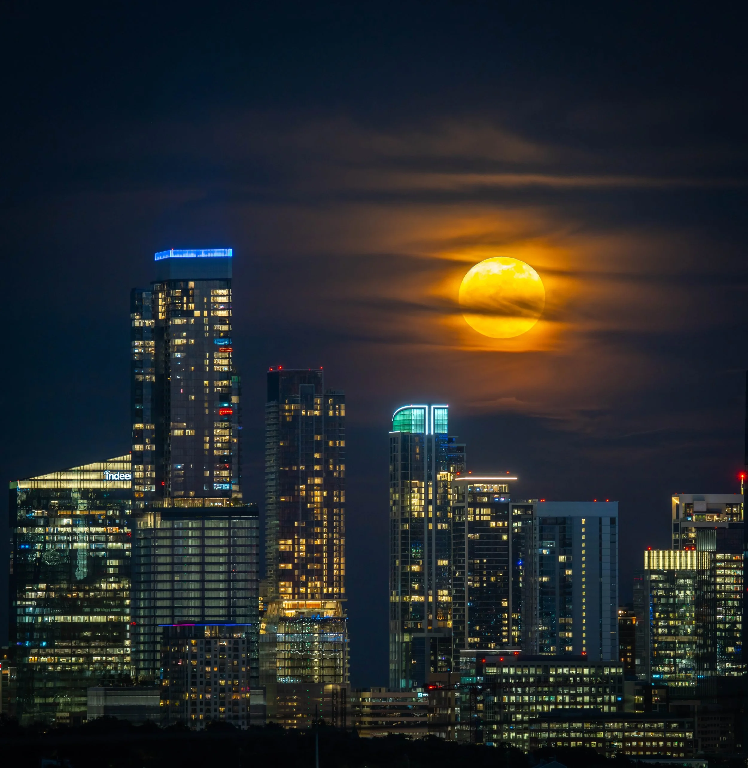 Nighttime city skyline with tall buildings illuminated and a large full moon in the sky, partly obscured by clouds.