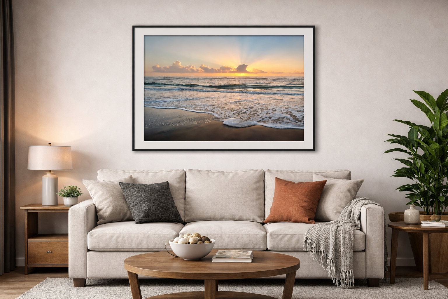 Living room with a beige sofa, gray and rust-colored pillows, a throw blanket, wooden side tables with a lamp and plants, a coffee table with decorative stones and books, and a framed beach sunset photograph on the wall.