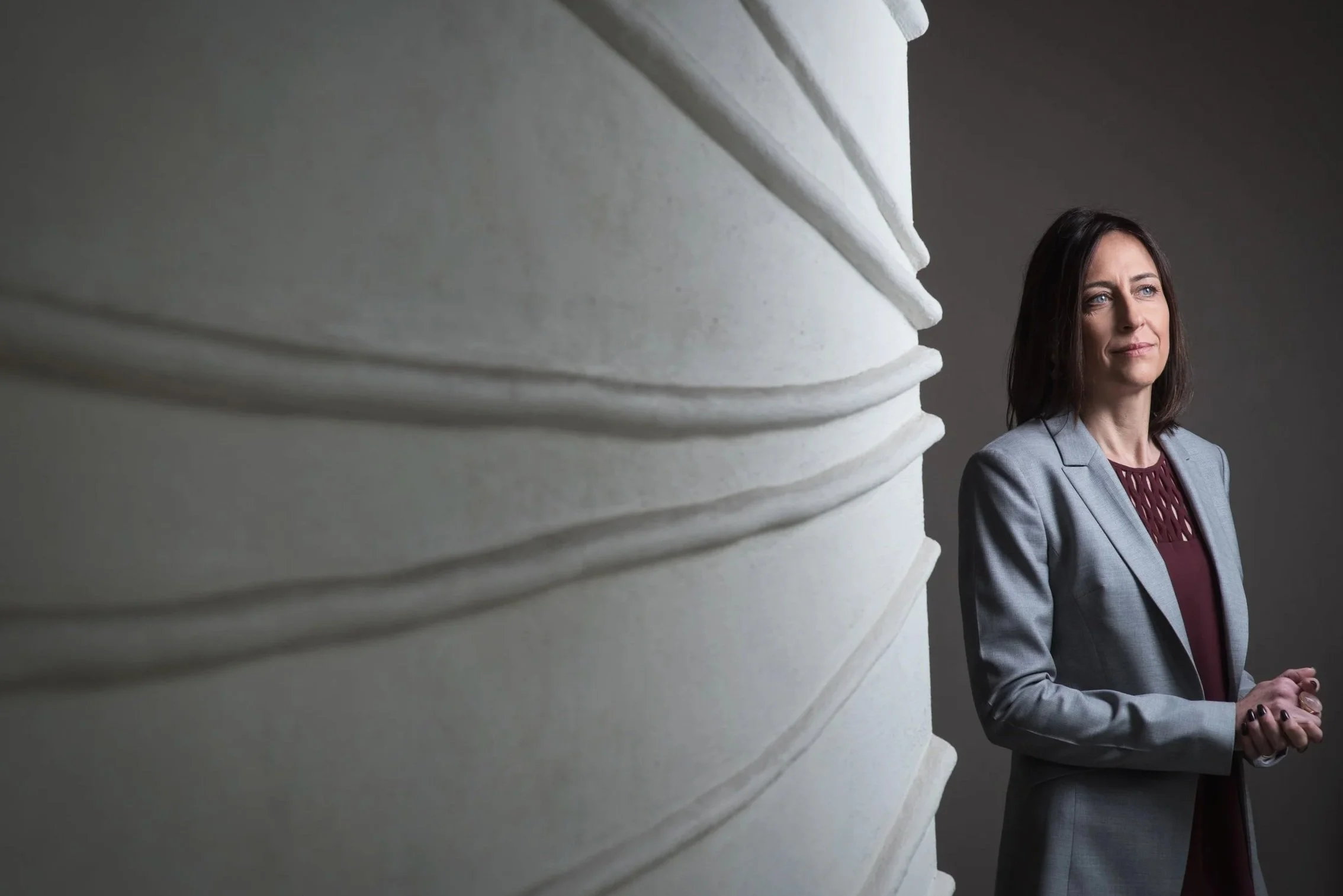Mulher de cabelo escuro e roupa social cinza, olhando pensativa para o lado, ao lado de uma grande escultura de pedra.