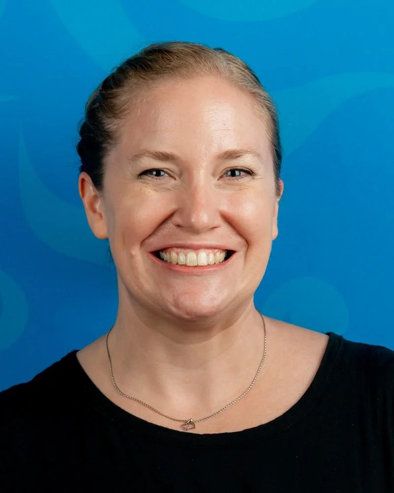 A woman with blonde hair tied back, smiling, wearing a black top and a silver necklace, against a blue background.