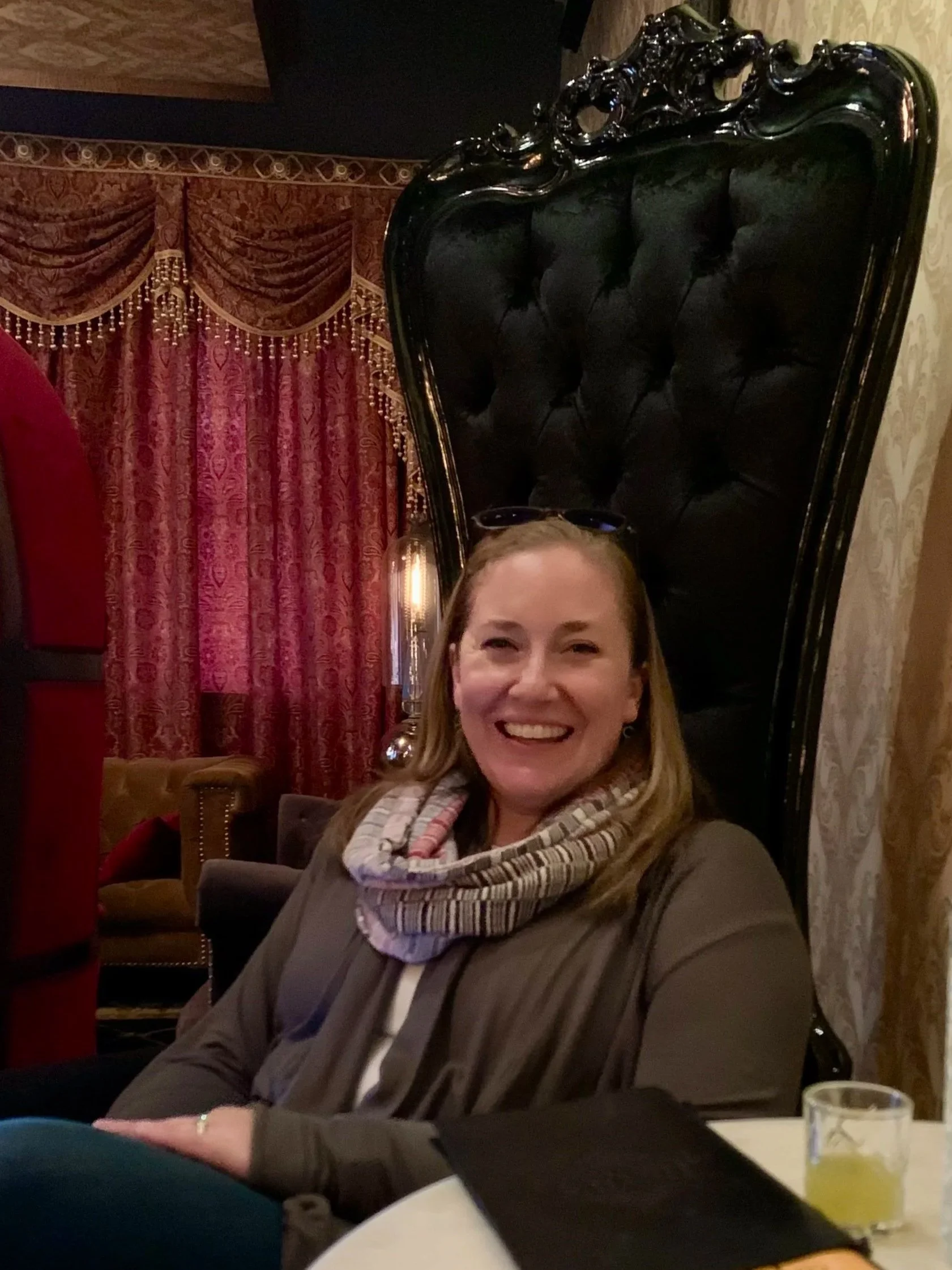 A woman with blonde hair smiling while sitting at a table in a luxurious room with ornate red and gold curtains. She is wearing a brown jacket and a striped scarf, with sunglasses on her head. There is a black high-backed chair behind her and a glass of yellow drink on the table.