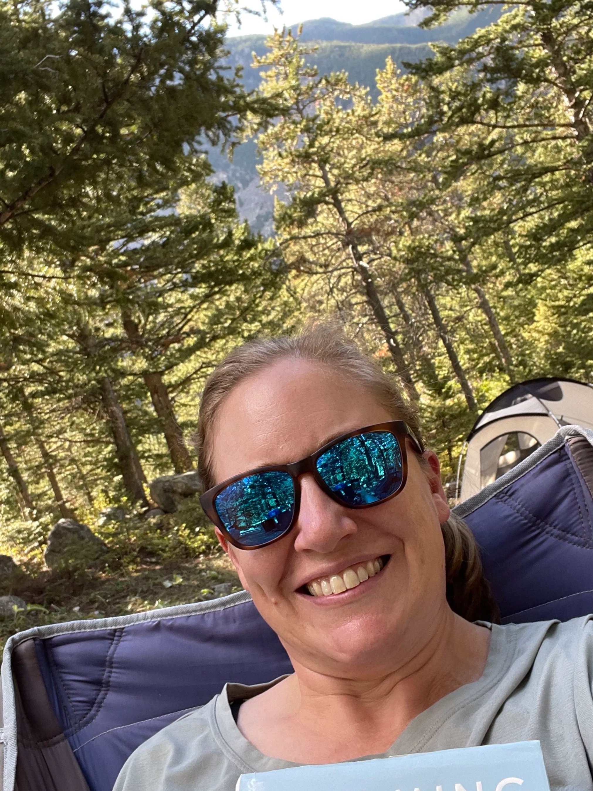 A woman smiling outdoors in a forest with trees and mountains in the background, wearing sunglasses and sitting in a camping chair.