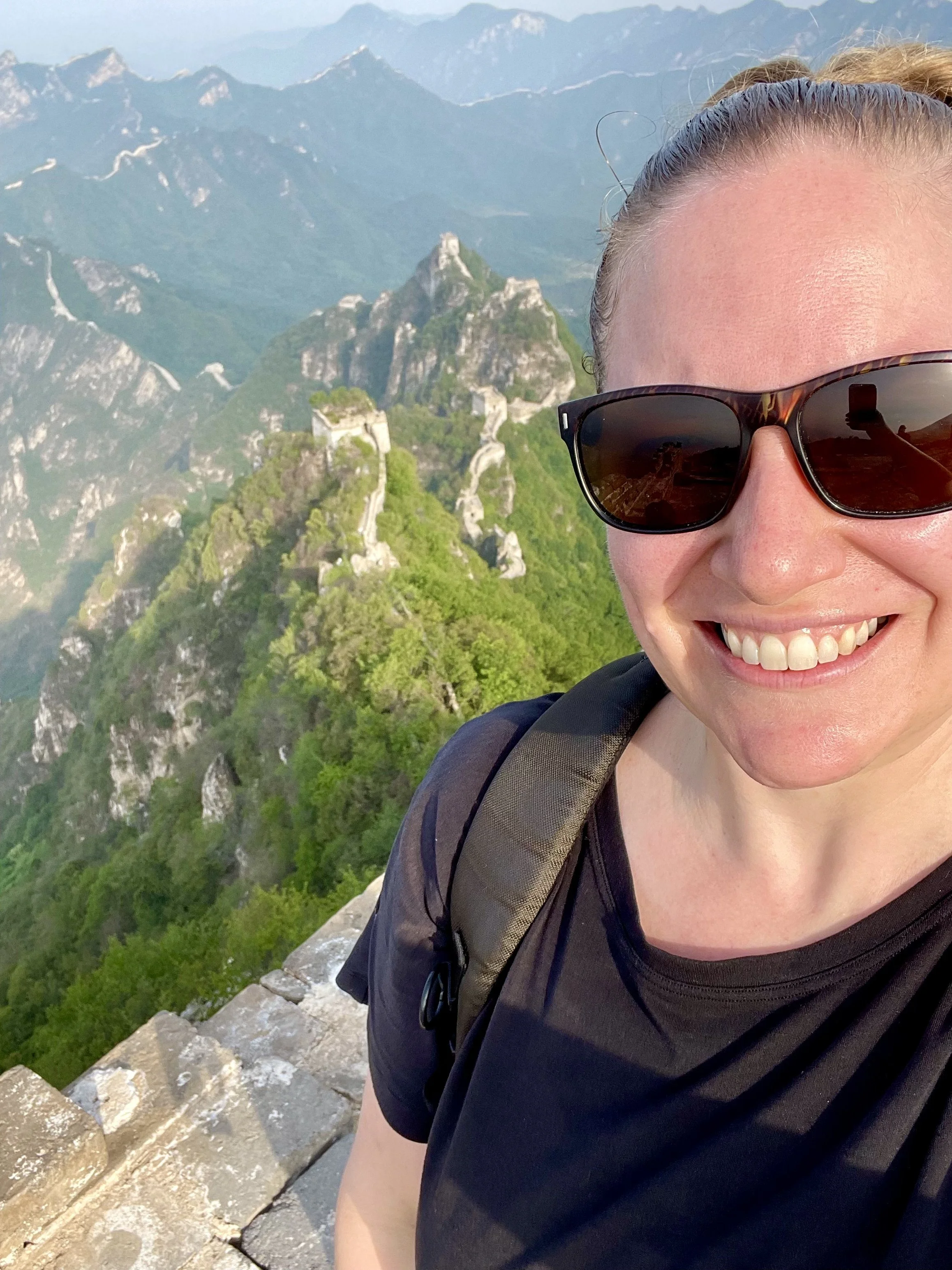 A woman taking a selfie at a mountain viewpoint with the Great Wall of China visible in the background, surrounded by lush green mountains.