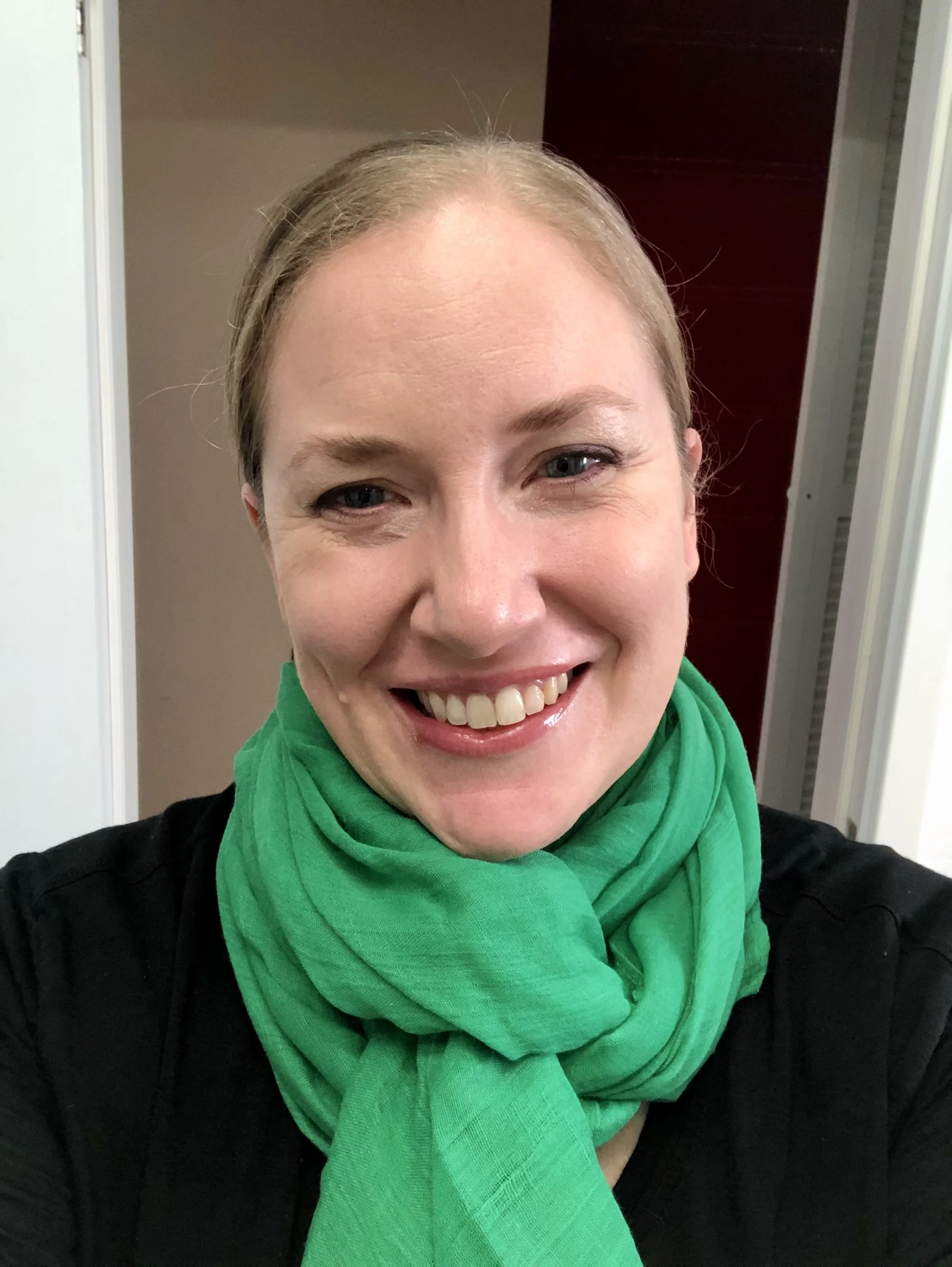 Close-up of a smiling woman with blonde hair, wearing a green scarf and black top. She is indoors with a neutral background.