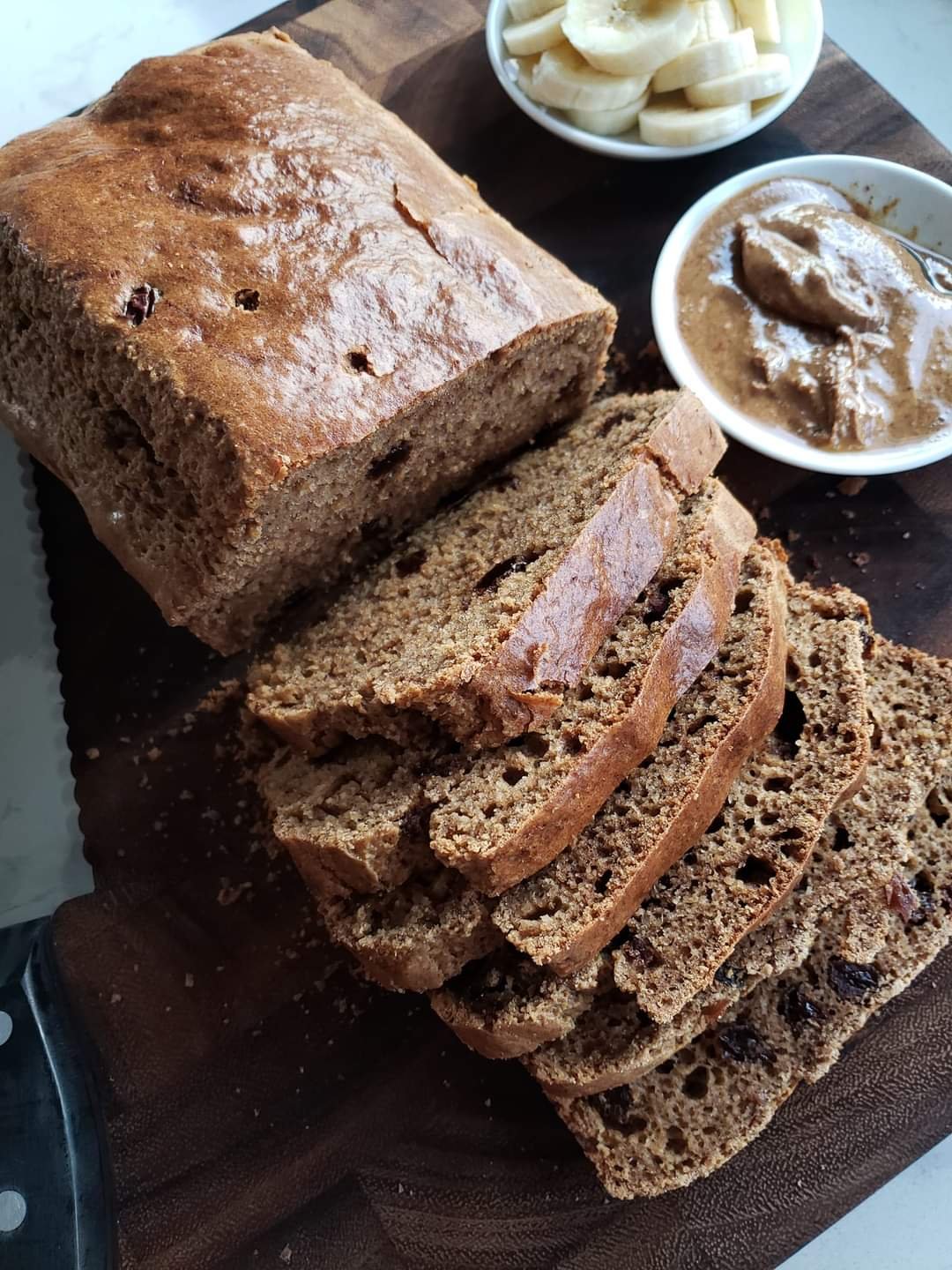 Slices of fruitcake on a wooden cutting board, with bowls of sliced bananas and a brown fruitcake frosting or spread nearby.