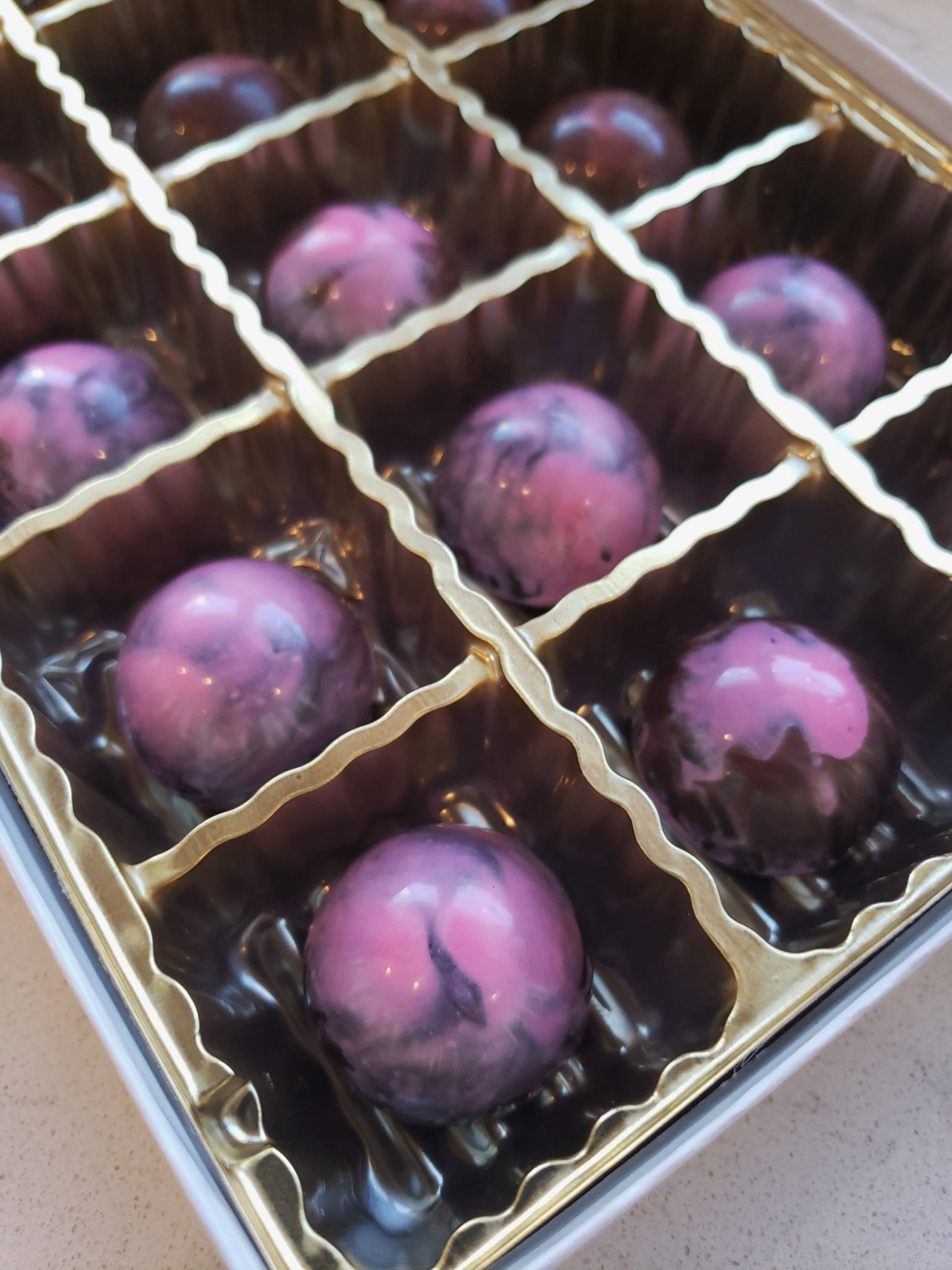 A golden box with individual compartments holding round purple and black chocolates or confections.