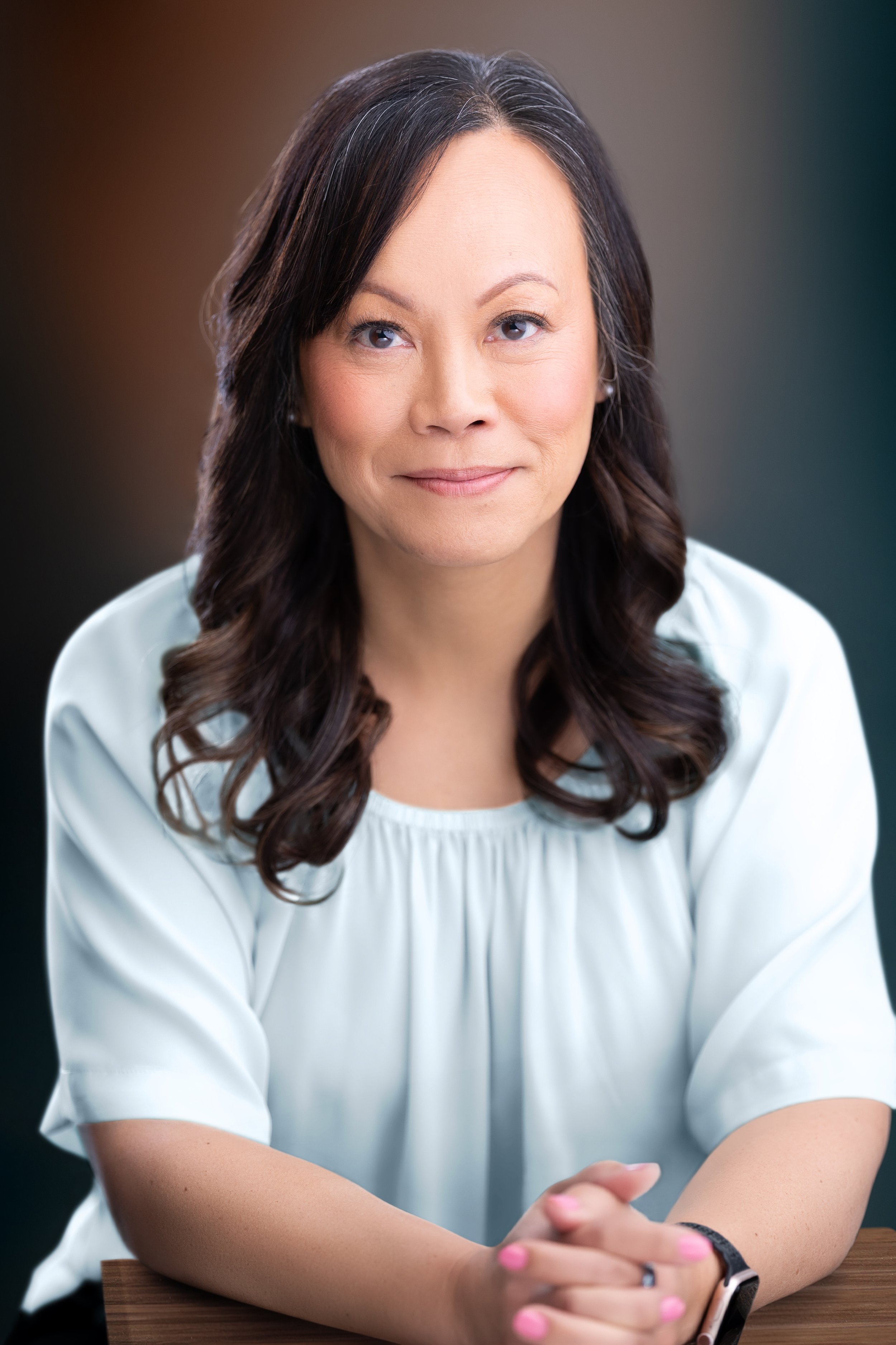 A woman with long dark wavy hair wearing a white blouse, sitting at a table with her hands clasped, looking slightly to the side with a gentle smile. Mediator at Liang Mediation & Consulting in Maryland.