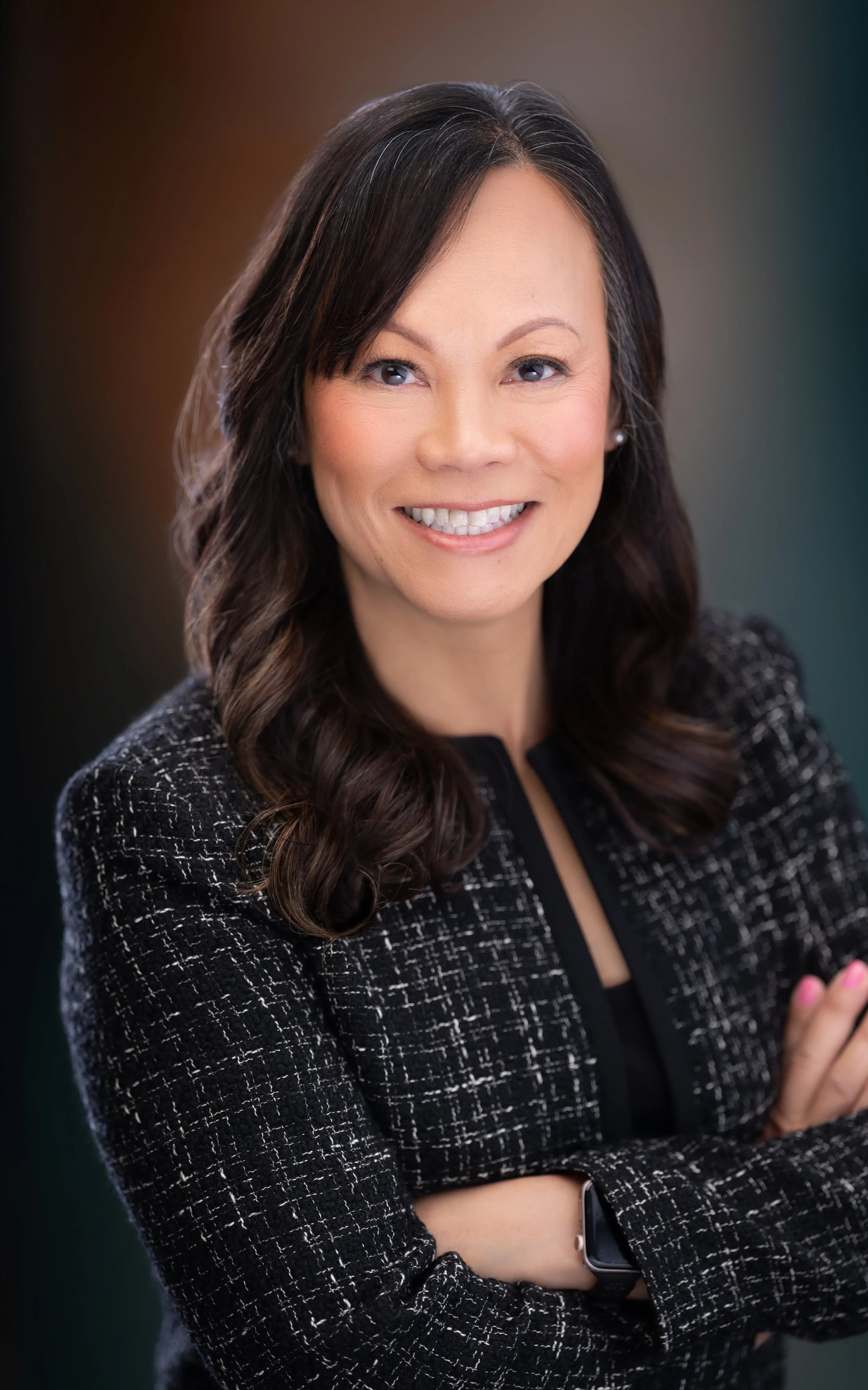 A smiling woman with dark wavy hair, wearing a black and white checked blazer and a smartwatch, standing with crossed arms against a blurred background. Mediator at Liang Mediation & Consulting in Maryland.